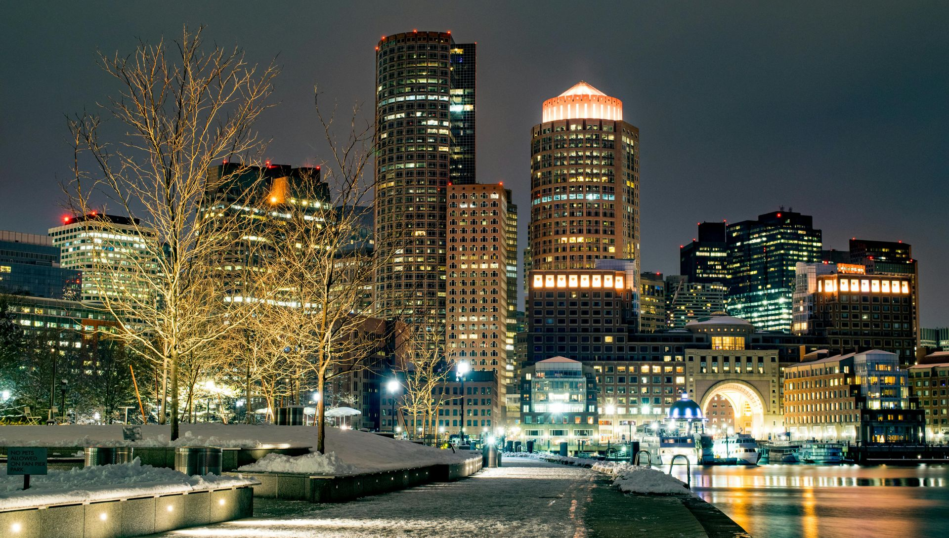 Night view of Boston skyline with illuminated skyscrapers, waterfront walkway, and snow-covered ground.