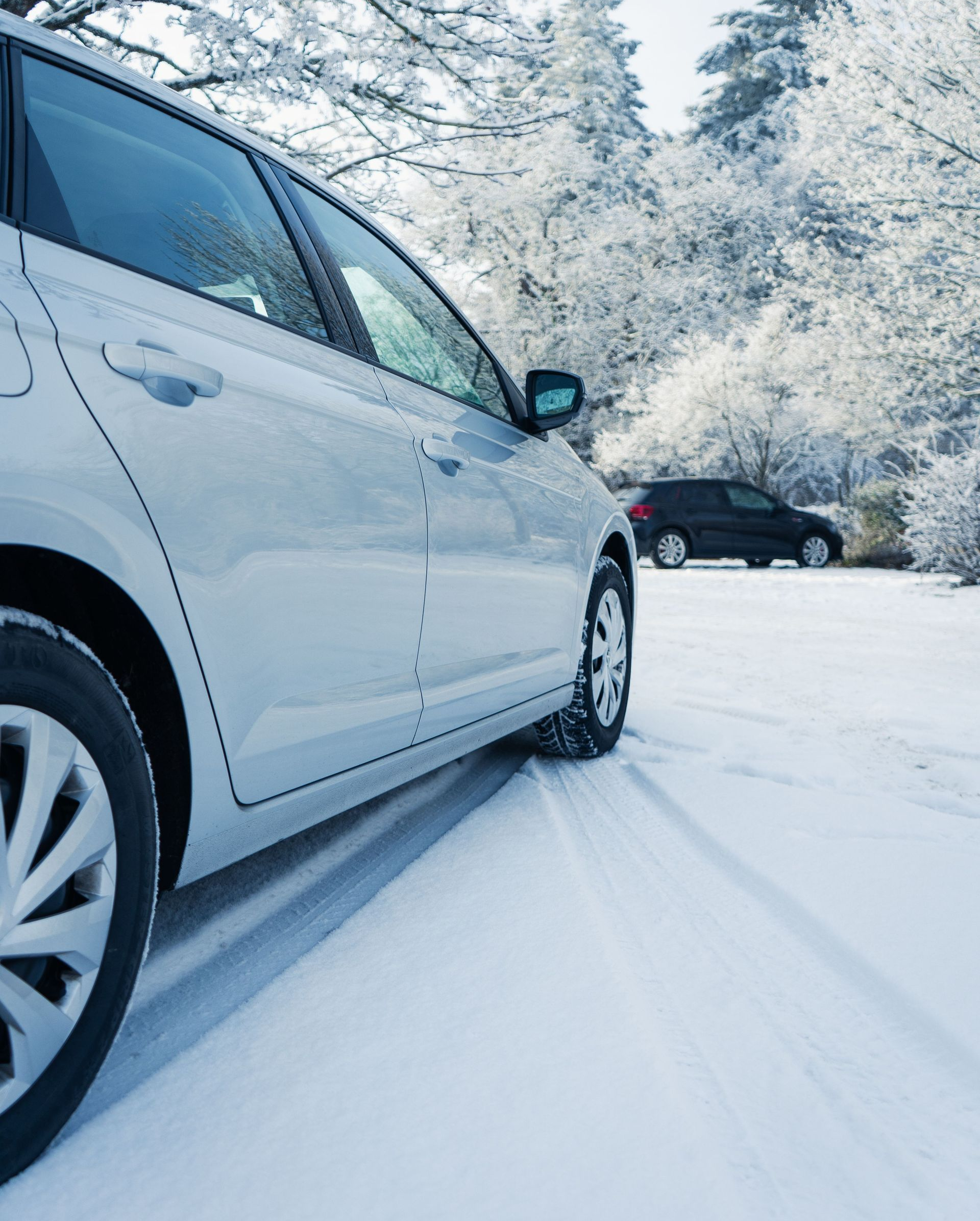 White SUV driving on a snowy road with tire tracks, winter trees and a dark car in the background