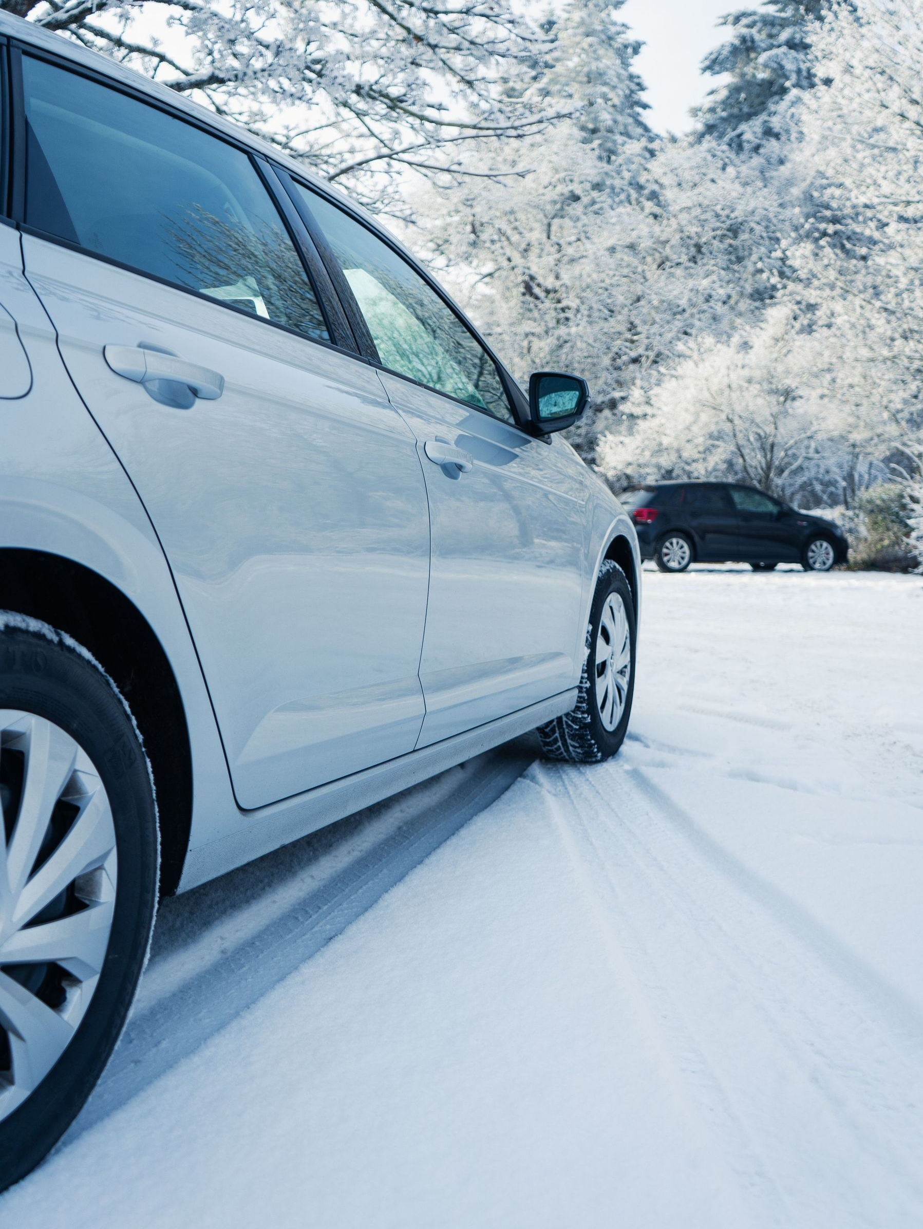 White SUV driving on a snowy road with tire tracks, winter trees and a dark car in the background