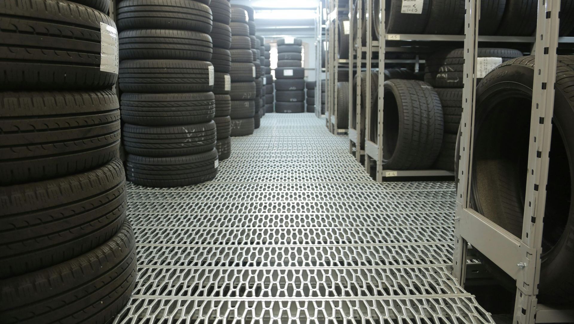 A storage room filled with stacks of car tires. Gray metal shelving lines the walls, and a grated floor covers the space.