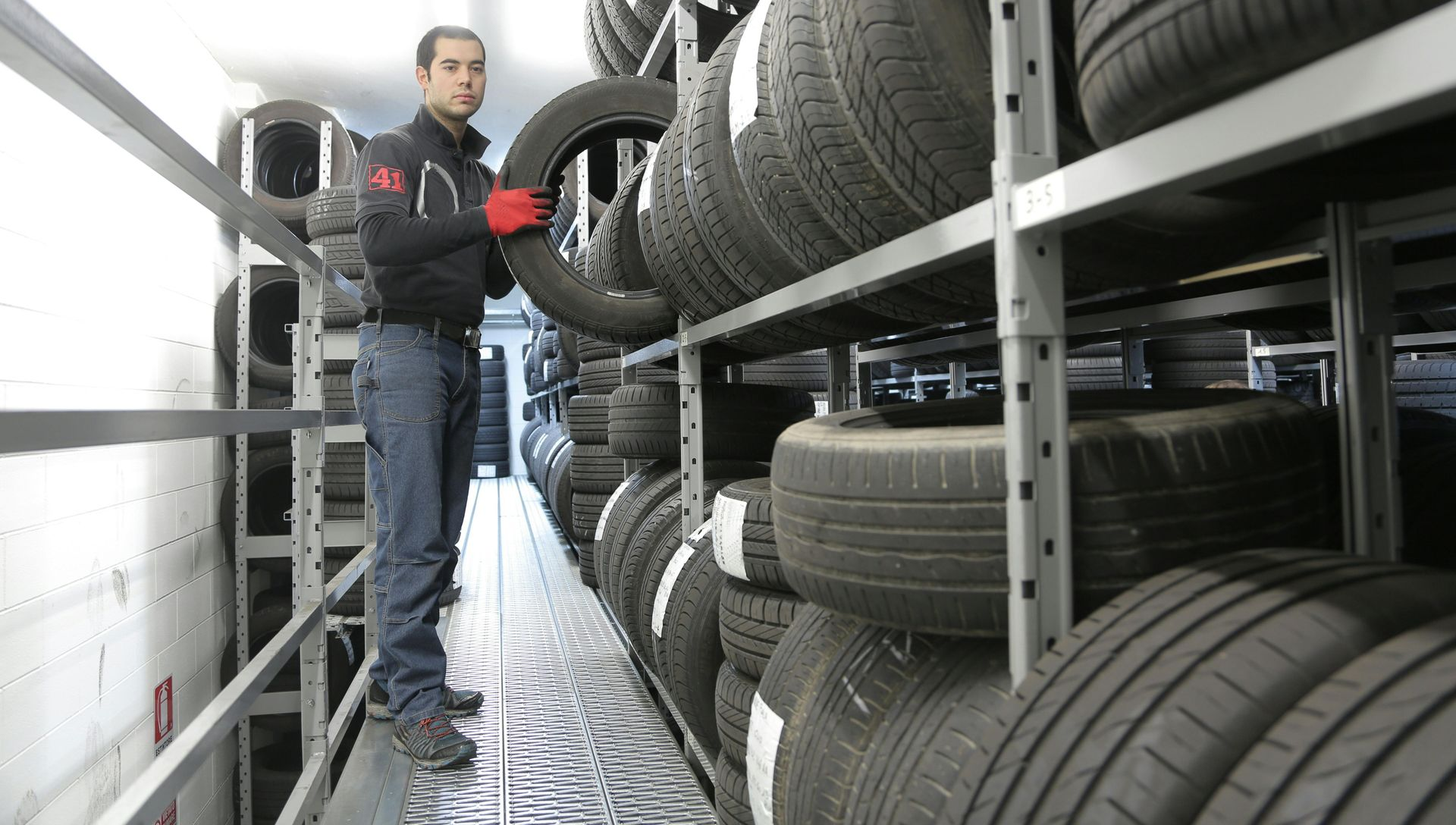 A warehouse worker in a dark uniform and red gloves selects a tire from a rack in a large storage facility.