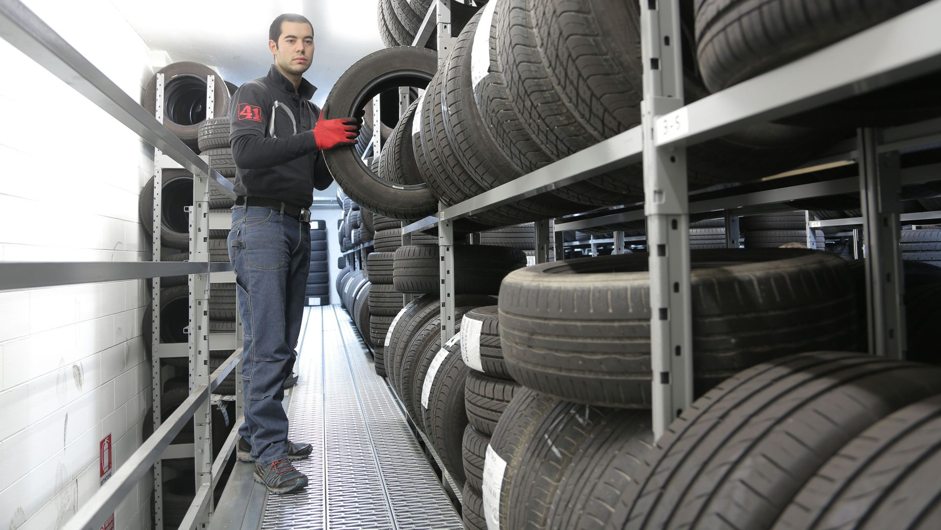 Mechanic inspecting stacked tires in a narrow storage aisle, wearing dark clothing and red gloves.