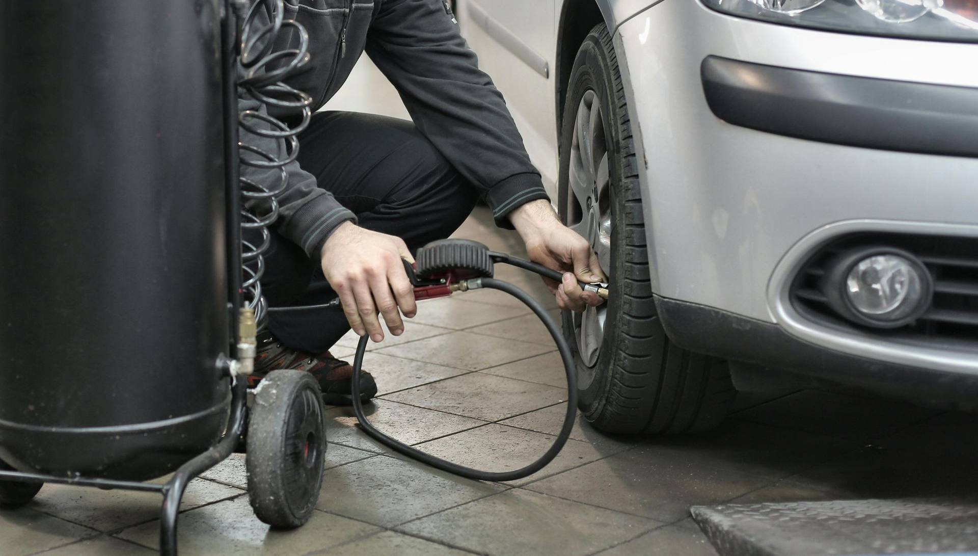 Person inflating car tire with air compressor in garage. Silver car, black equipment.