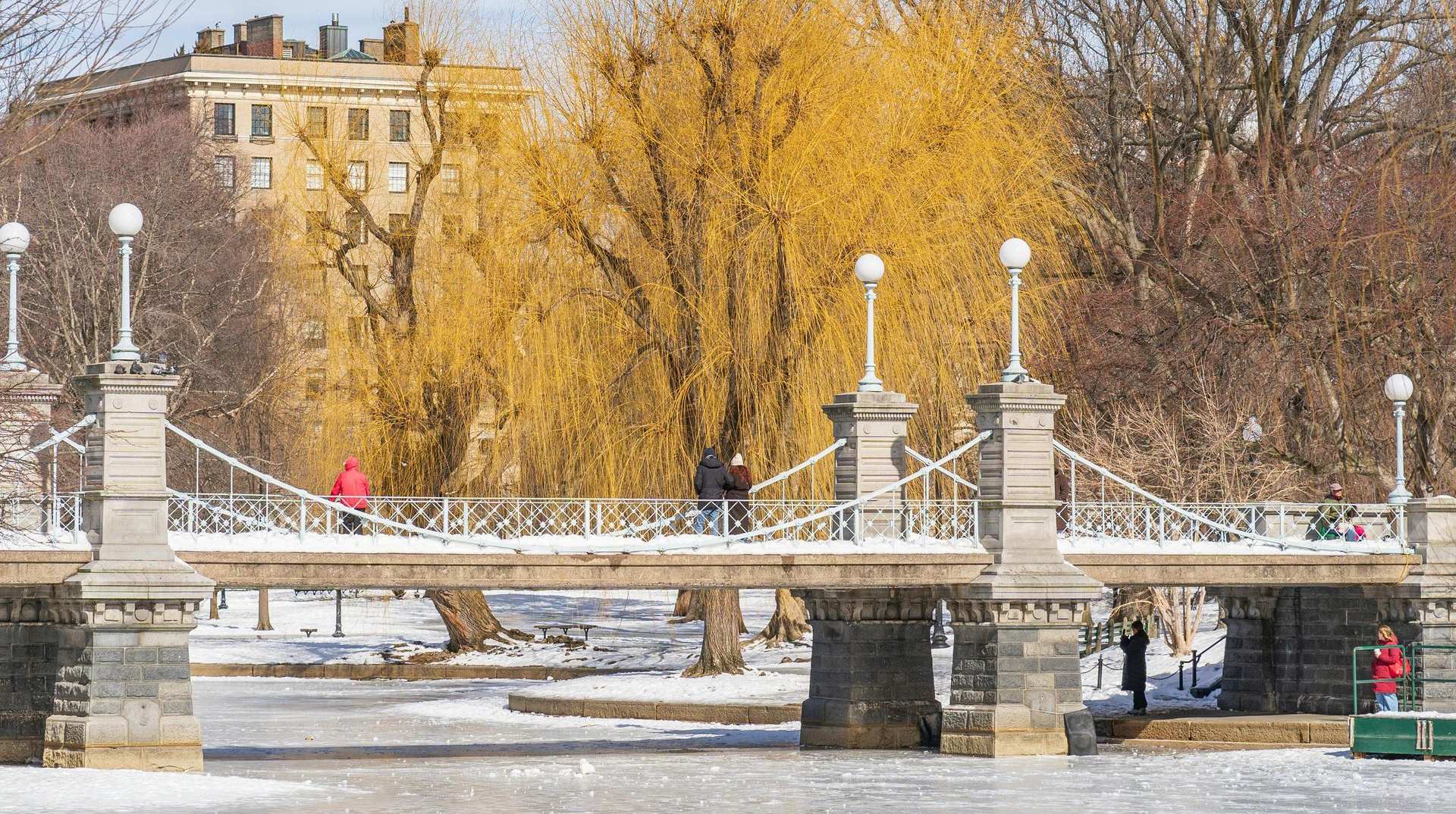 Bridge over frozen water in Boston with people walking. Buildings and yellow trees in background.