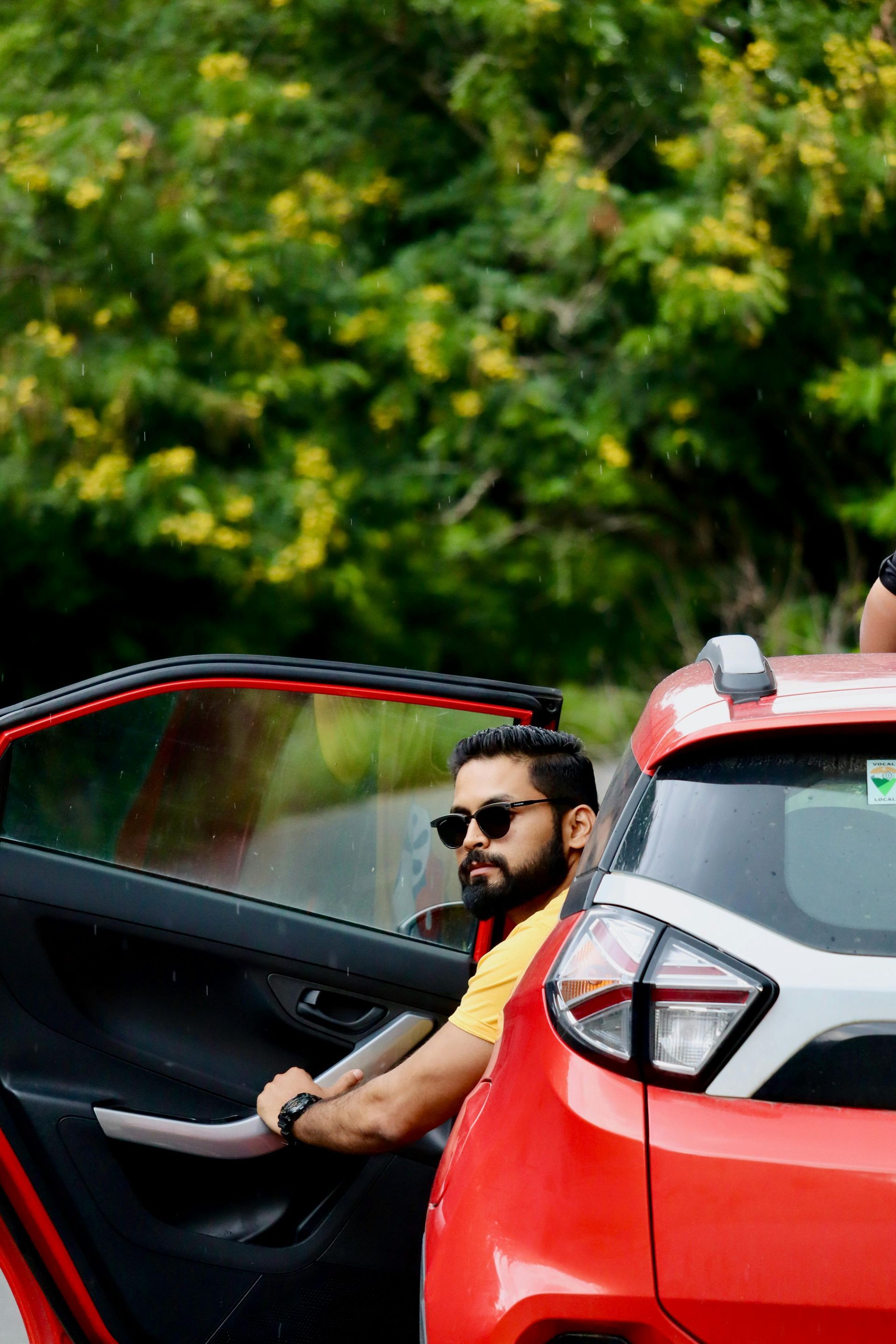 Man sitting in an open red car door, wearing sunglasses, with green foliage in the background