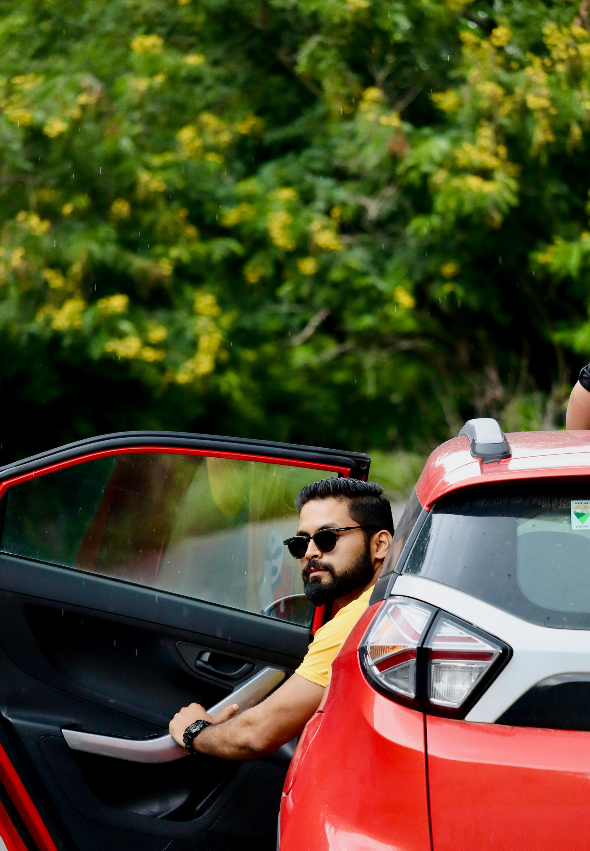 Man sitting in an open red car door, wearing sunglasses, with green foliage in the background