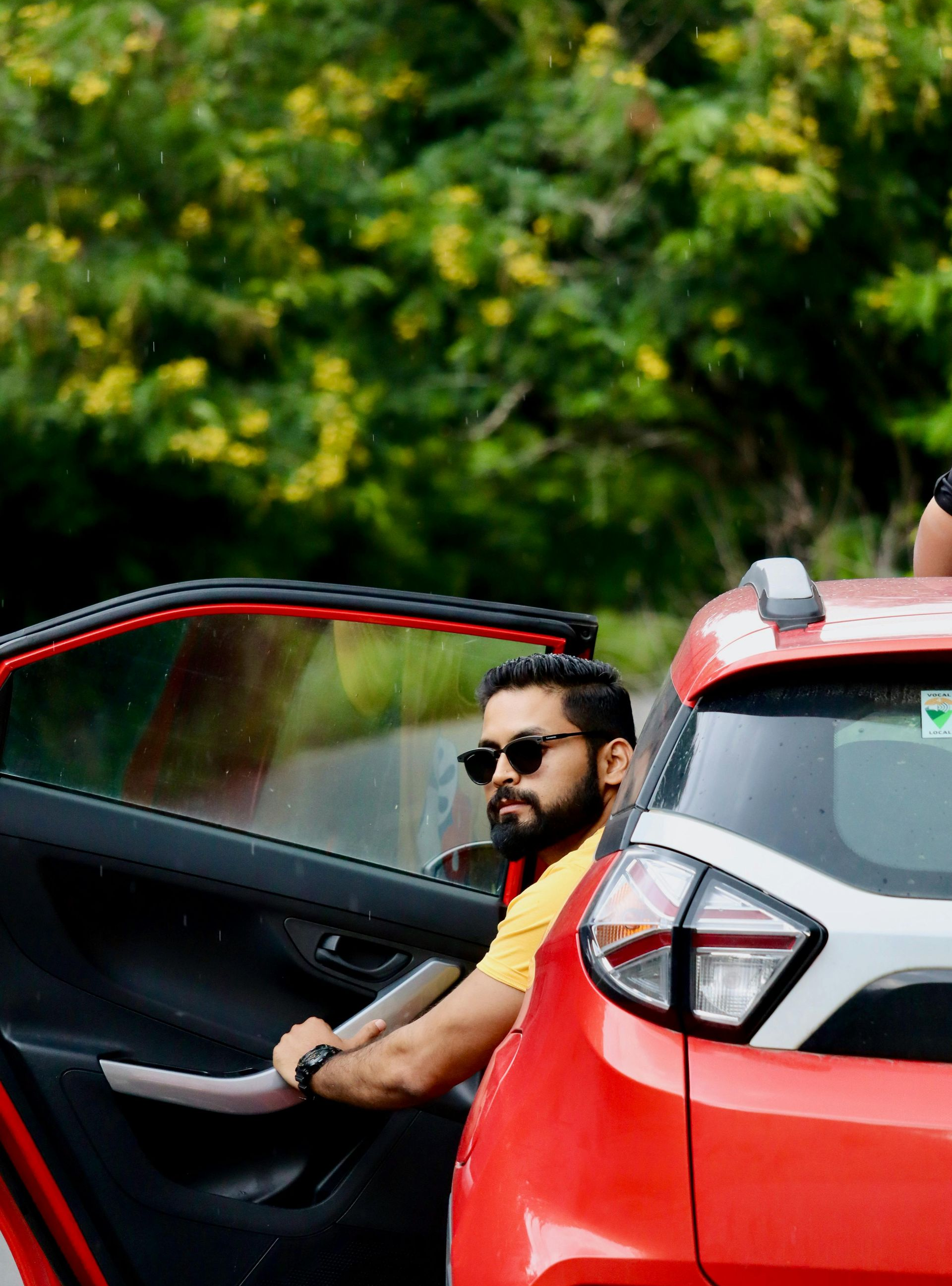 Man sitting in an open red car door, wearing sunglasses, with green foliage in the background