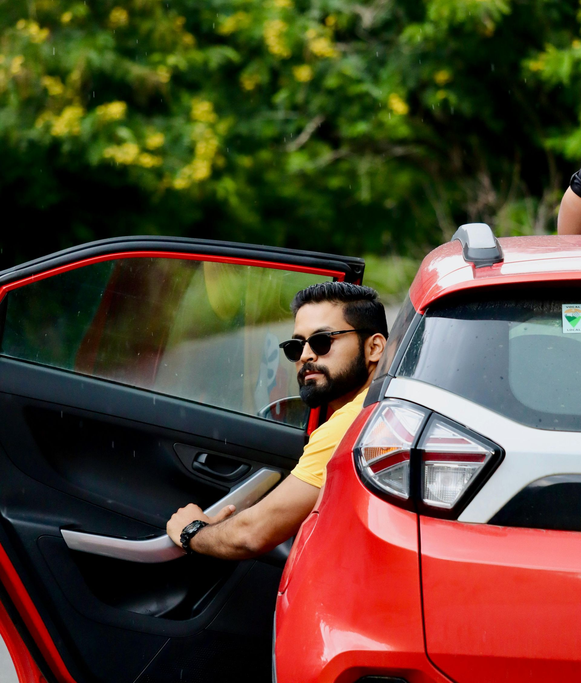 Man sitting in an open red car door, wearing sunglasses, with green foliage in the background