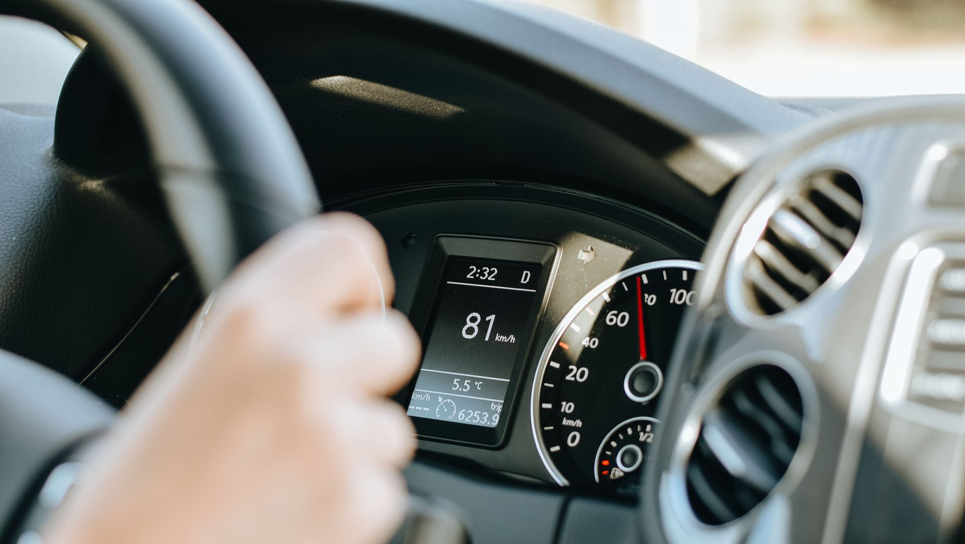 Driver holding steering wheel inside a car, with dashboard speedometer and air vent visible.