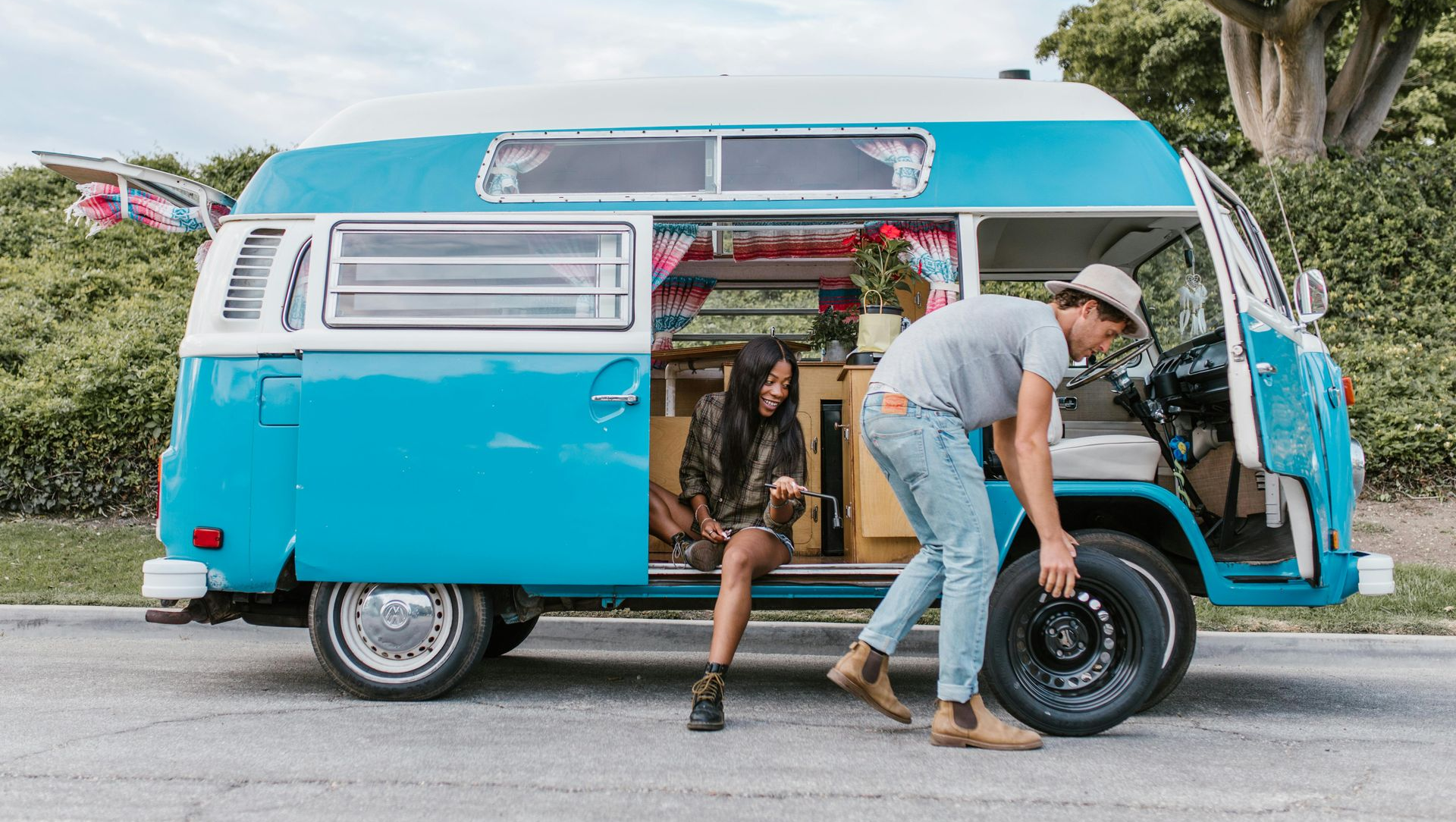 Man changing a tire on a blue and white vintage van; woman sits inside, looking out.