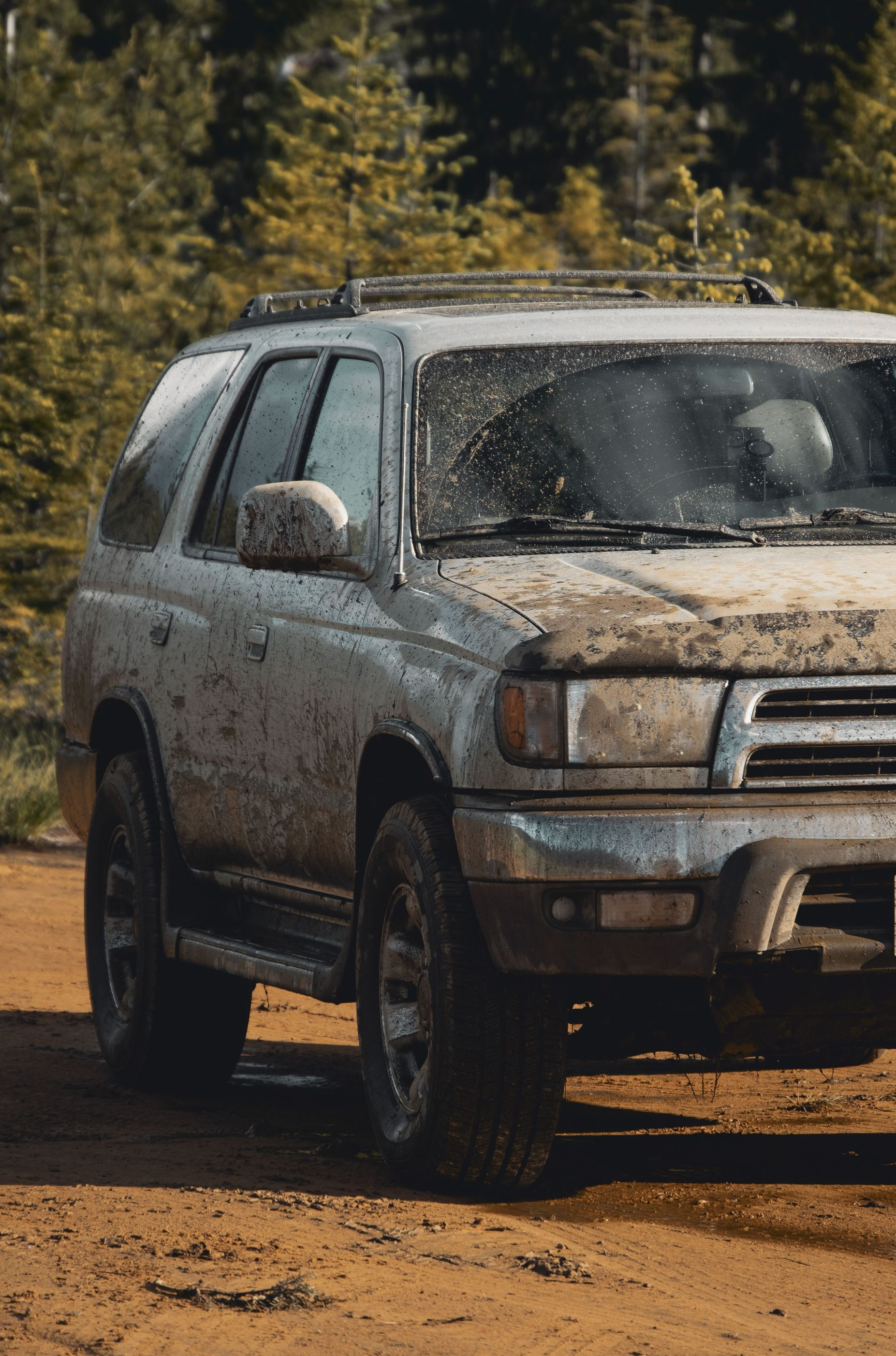Mud-covered silver SUV parked on a dirt road with trees in the background
