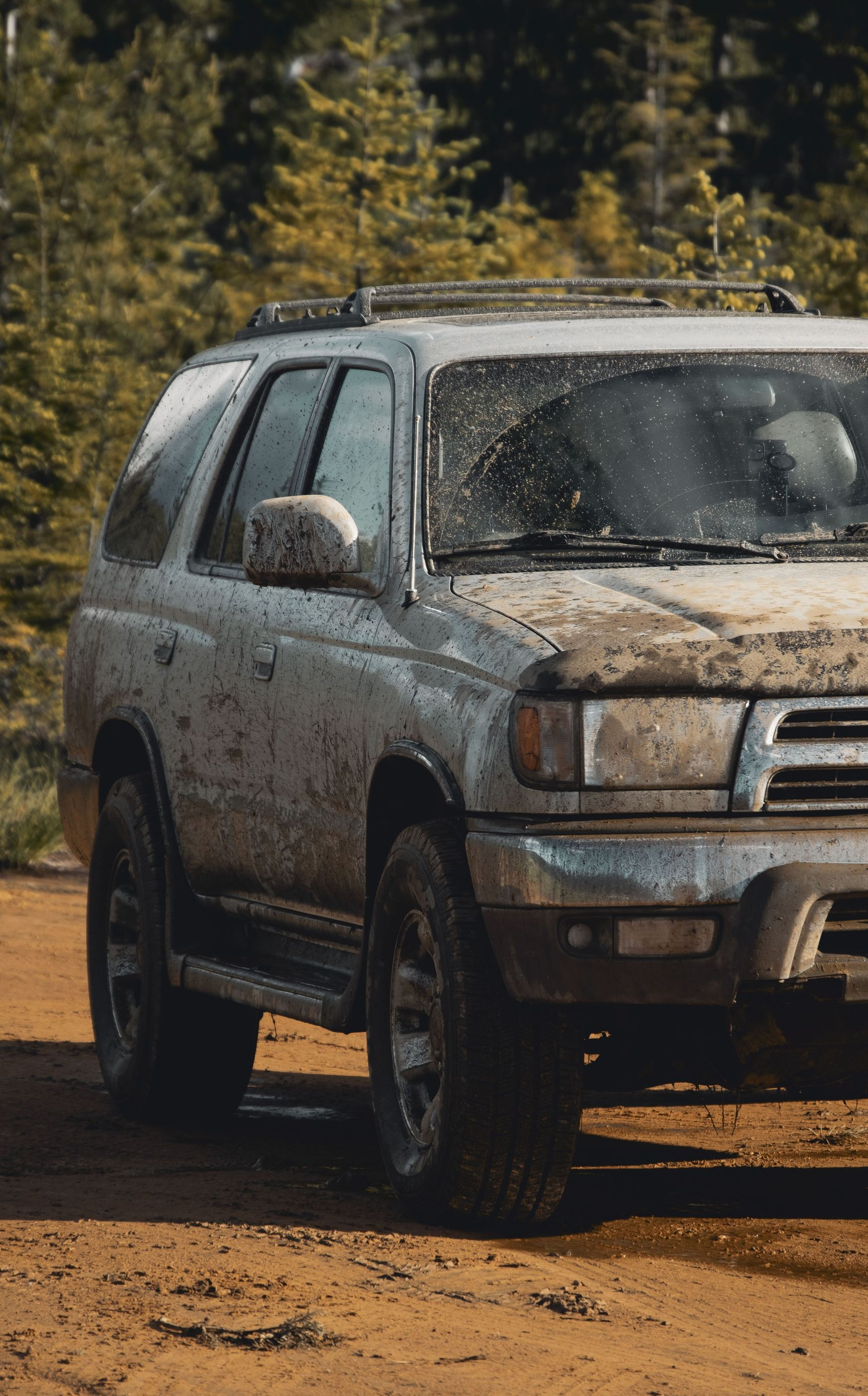 Mud-covered silver SUV parked on a dirt road with trees in the background