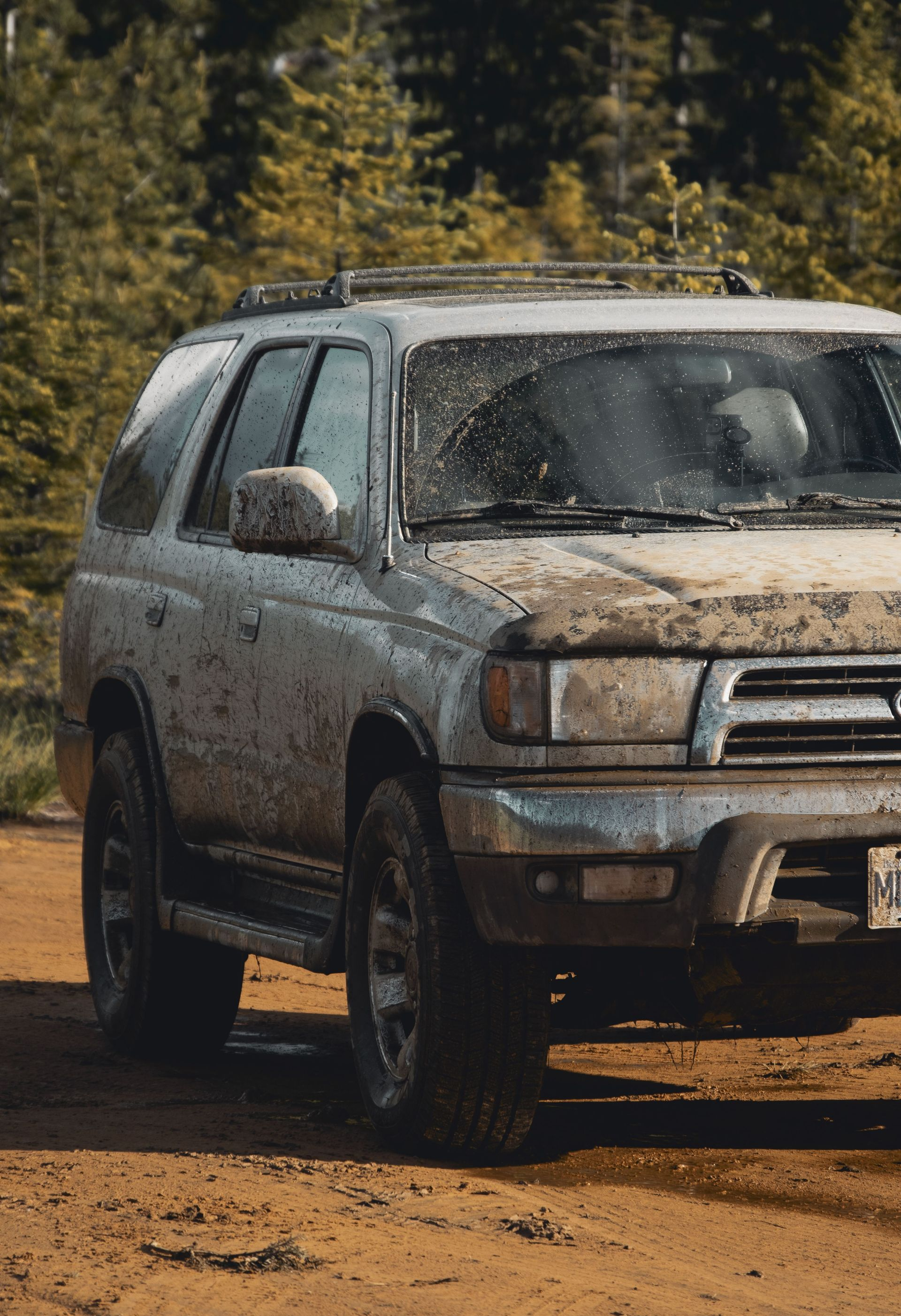 Mud-covered silver SUV parked on a dirt road with trees in the background