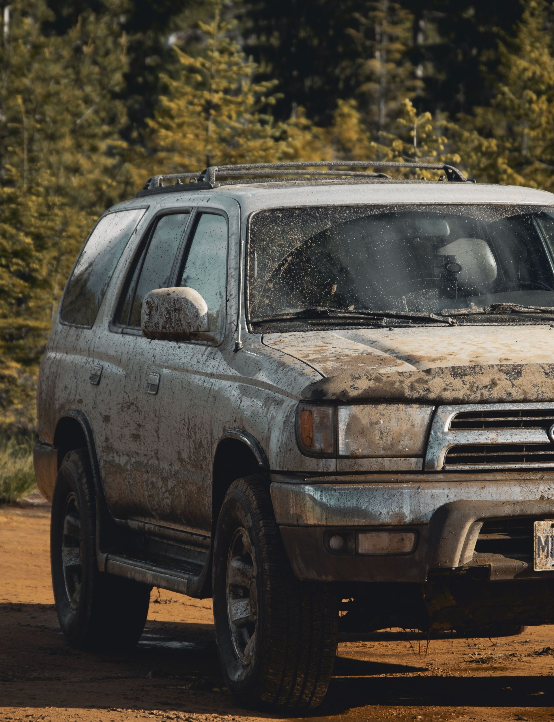 Mud-covered silver SUV parked on a dirt road with trees in the background
