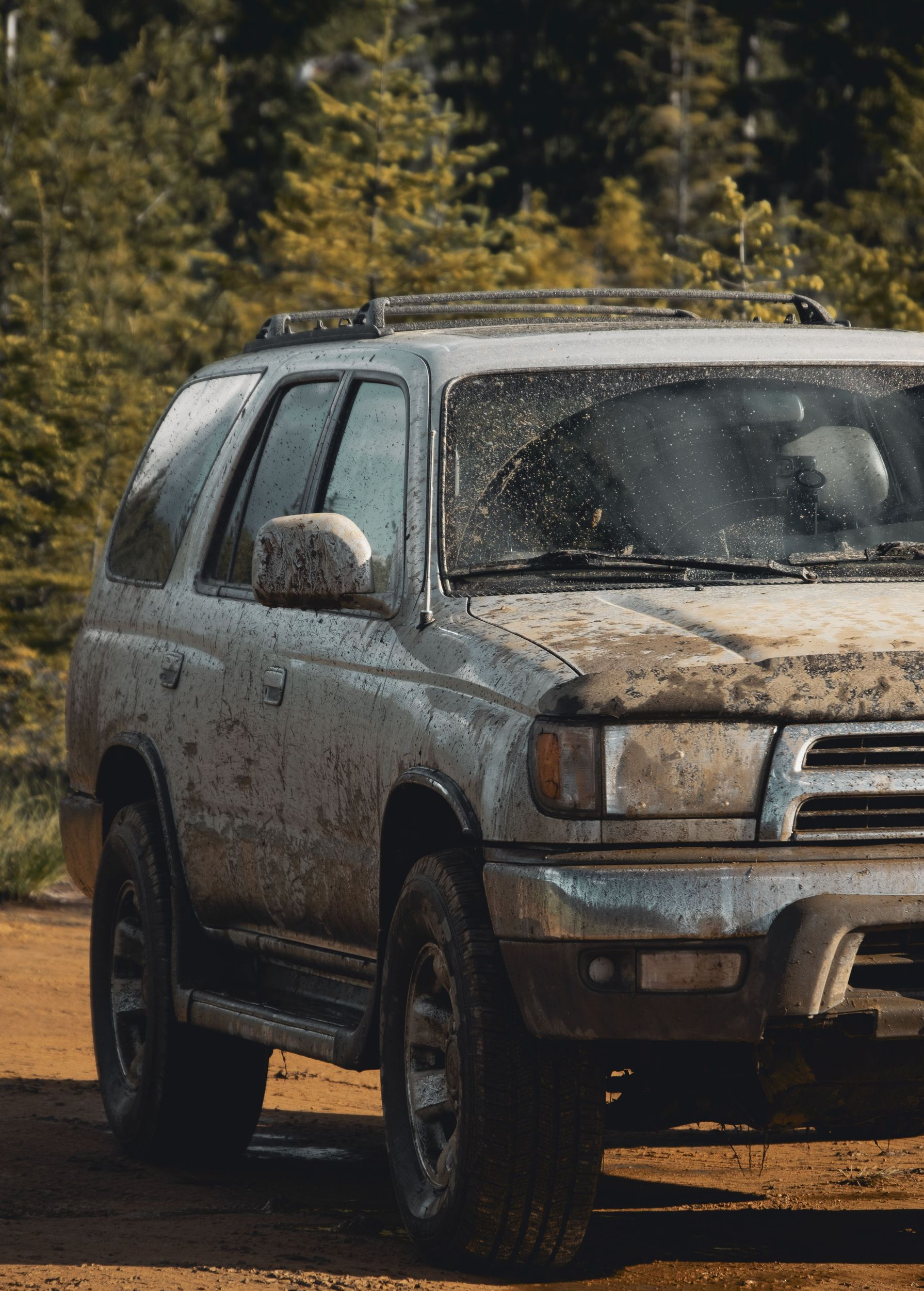 Mud-covered silver SUV parked on a dirt road with trees in the background