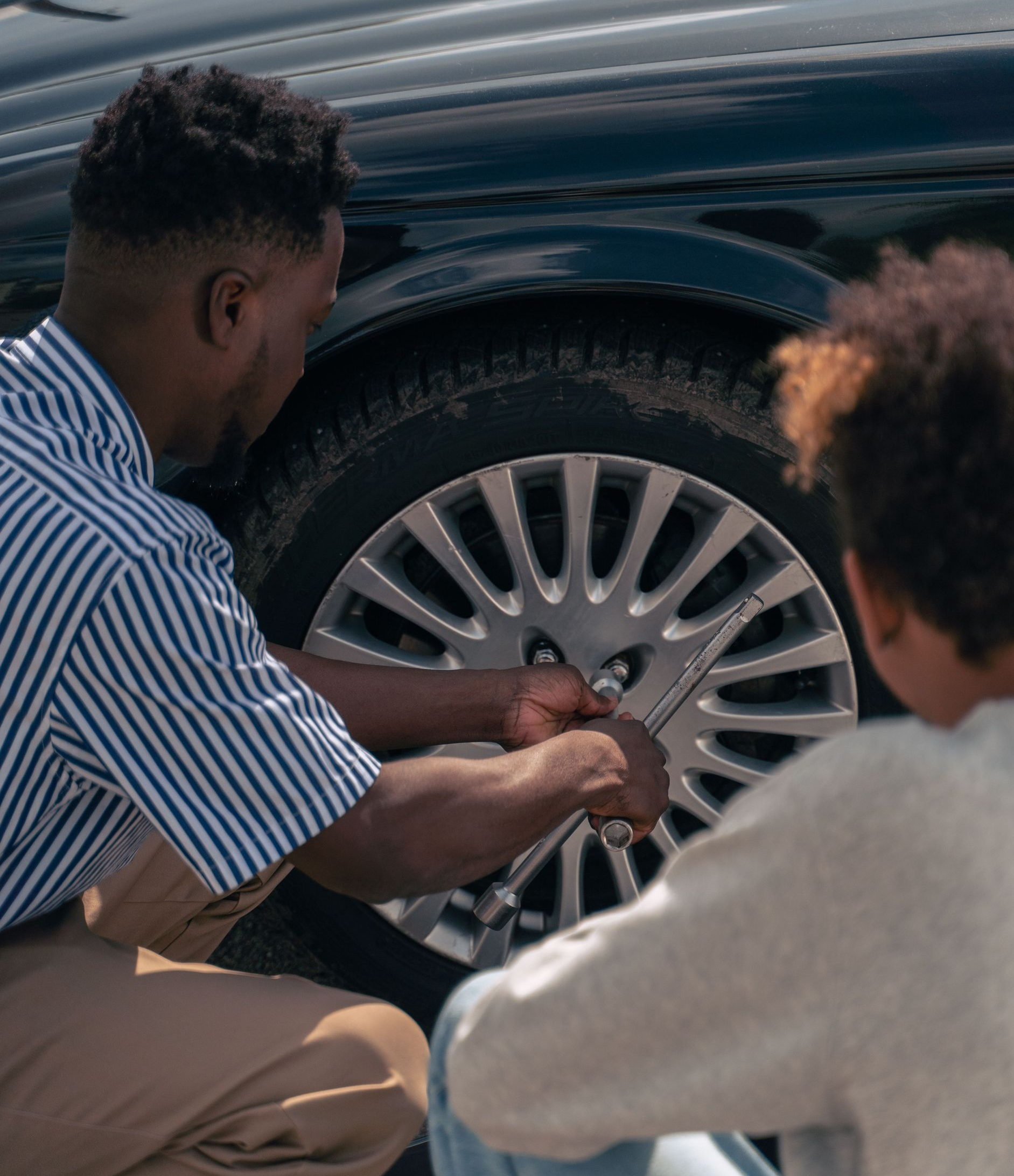 Two people crouch by a car wheel, tightening a lug nut with a wrench.