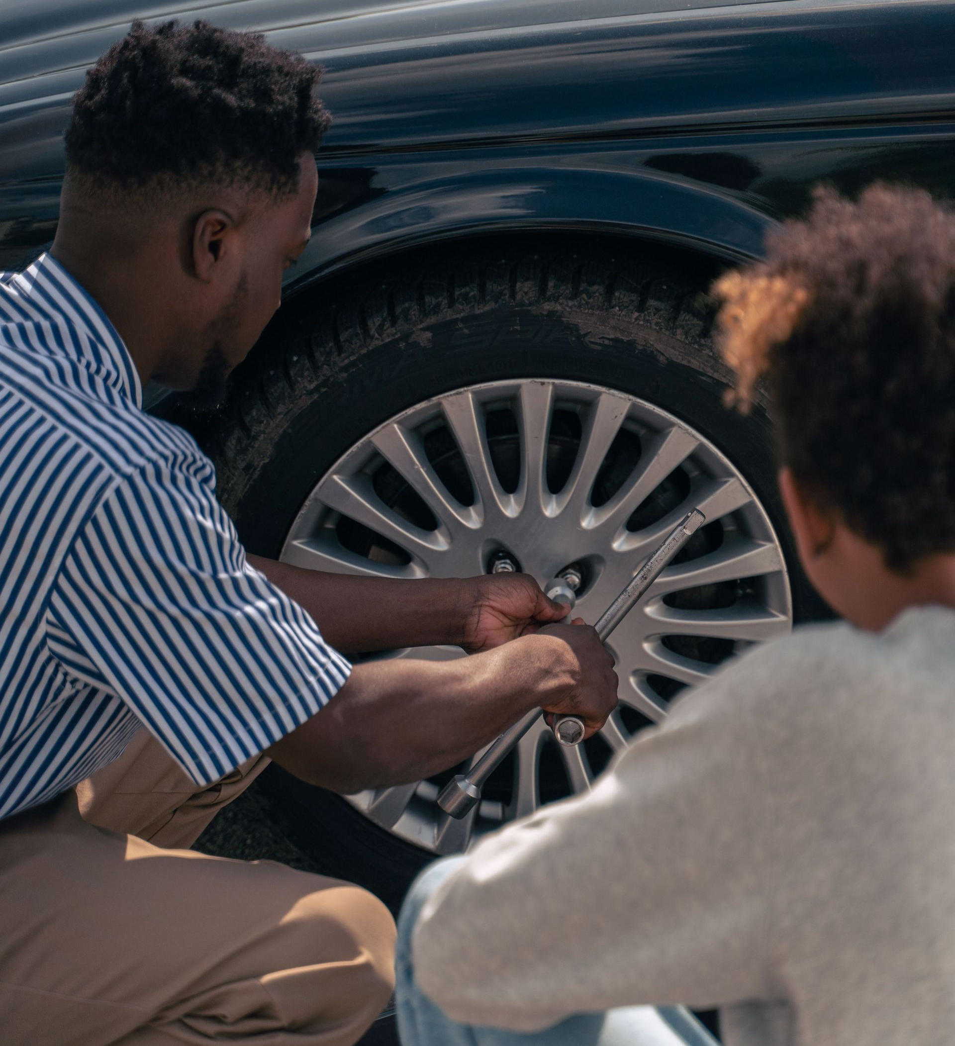 Two people crouch by a car wheel, tightening a lug nut with a wrench.