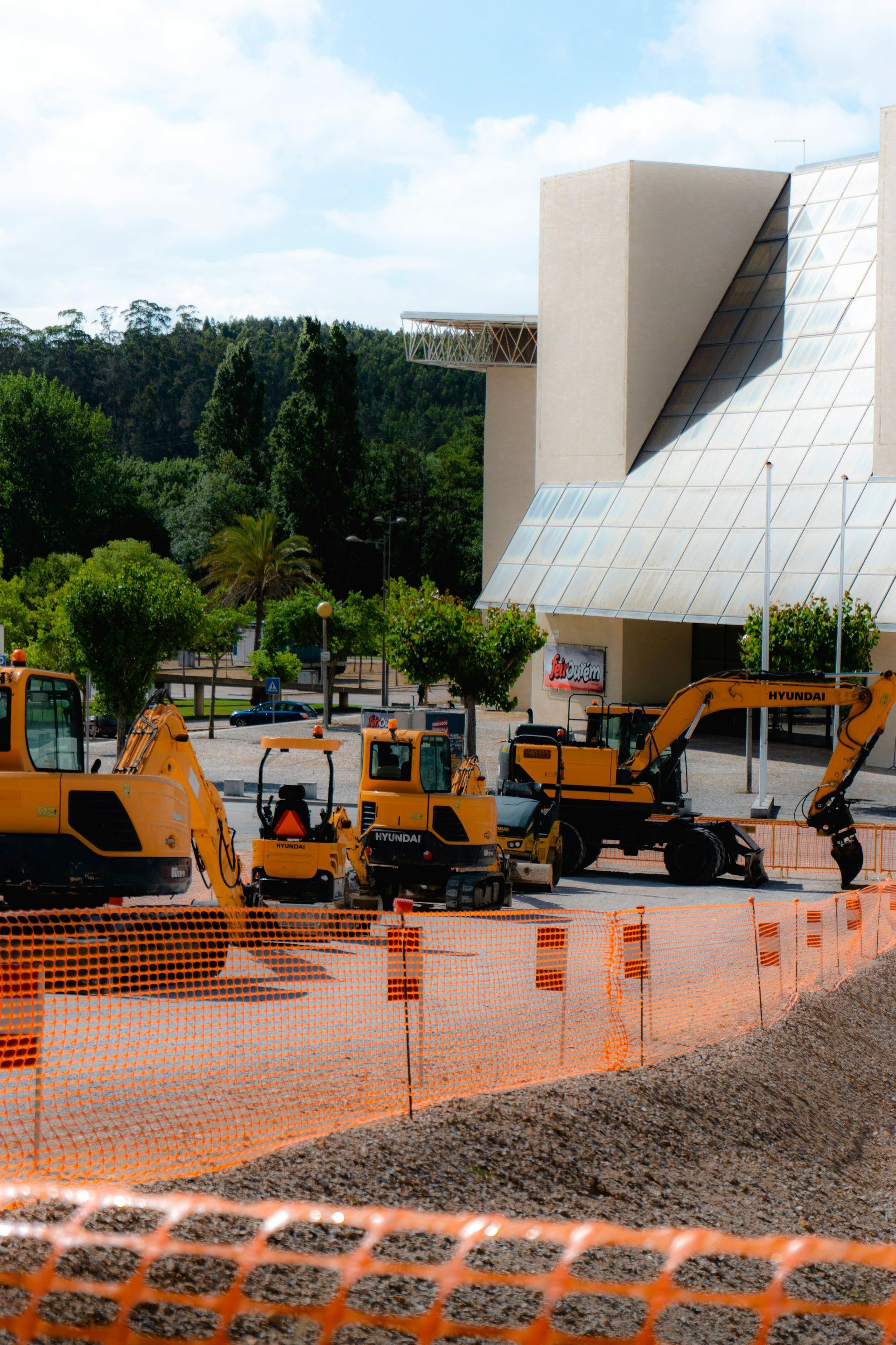 Construction site with yellow excavators behind orange safety fencing beside a modern white building