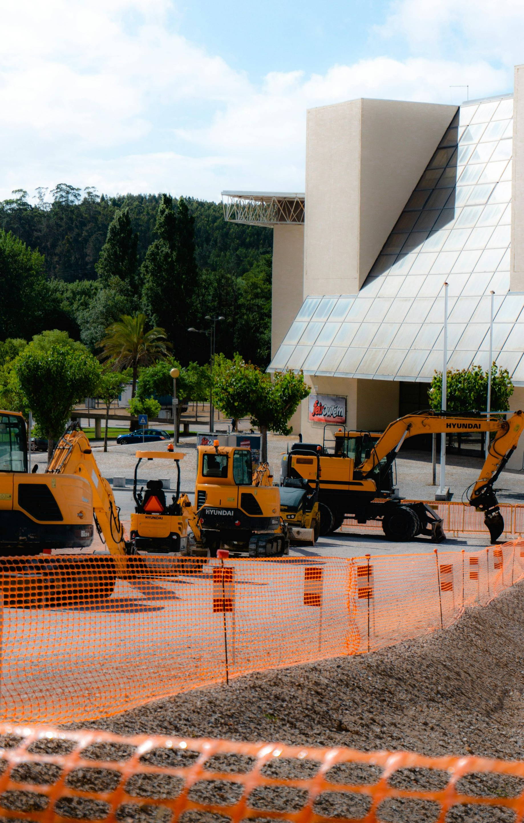 Construction site with yellow excavators behind orange safety fencing beside a modern white building