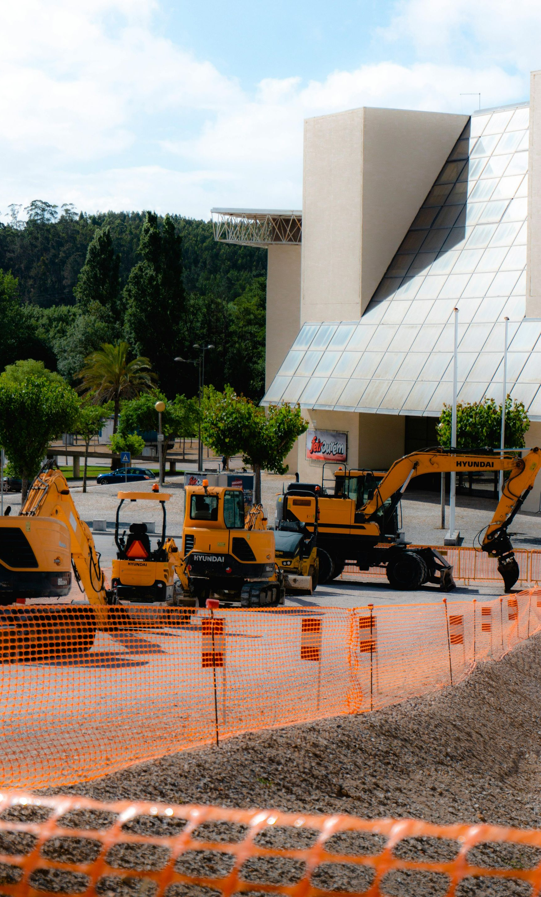 Construction site with yellow excavators behind orange safety fencing beside a modern white building