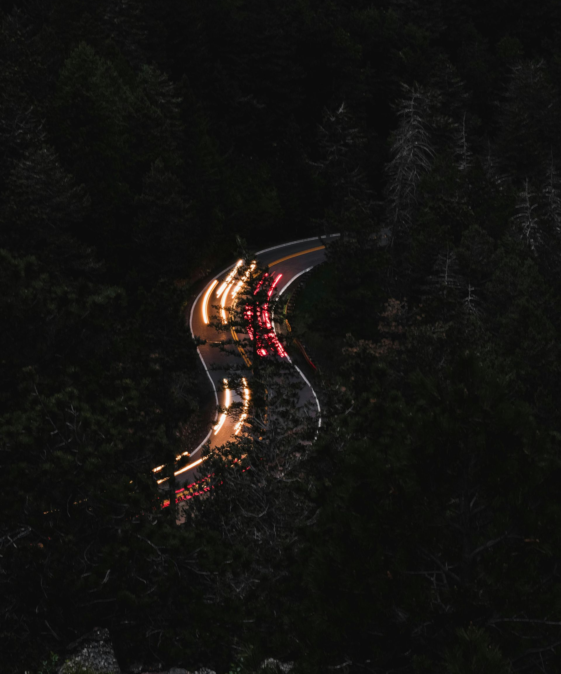 Nighttime aerial view of a winding road with blurred car lights snaking through a dark forest.