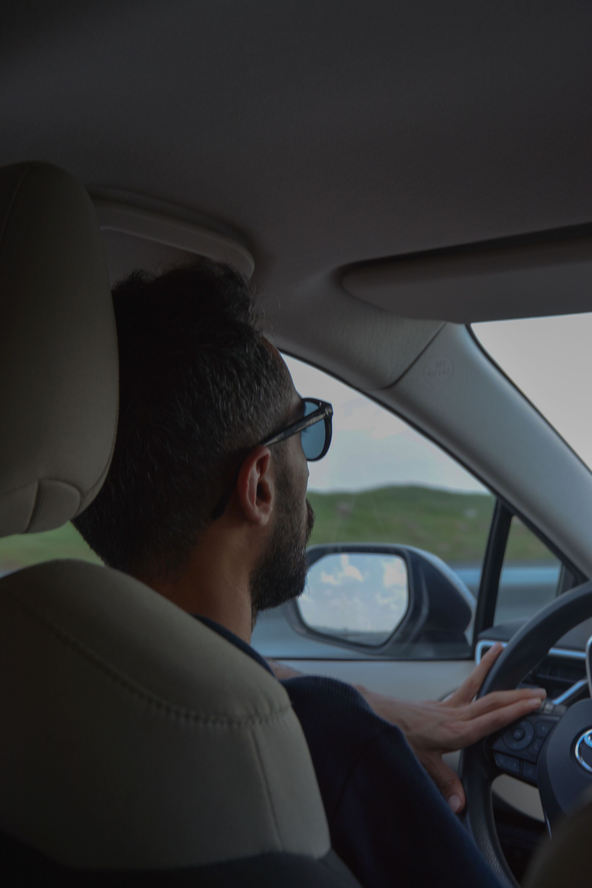 Driver wearing glasses steering a car on a road, viewed from the back seat