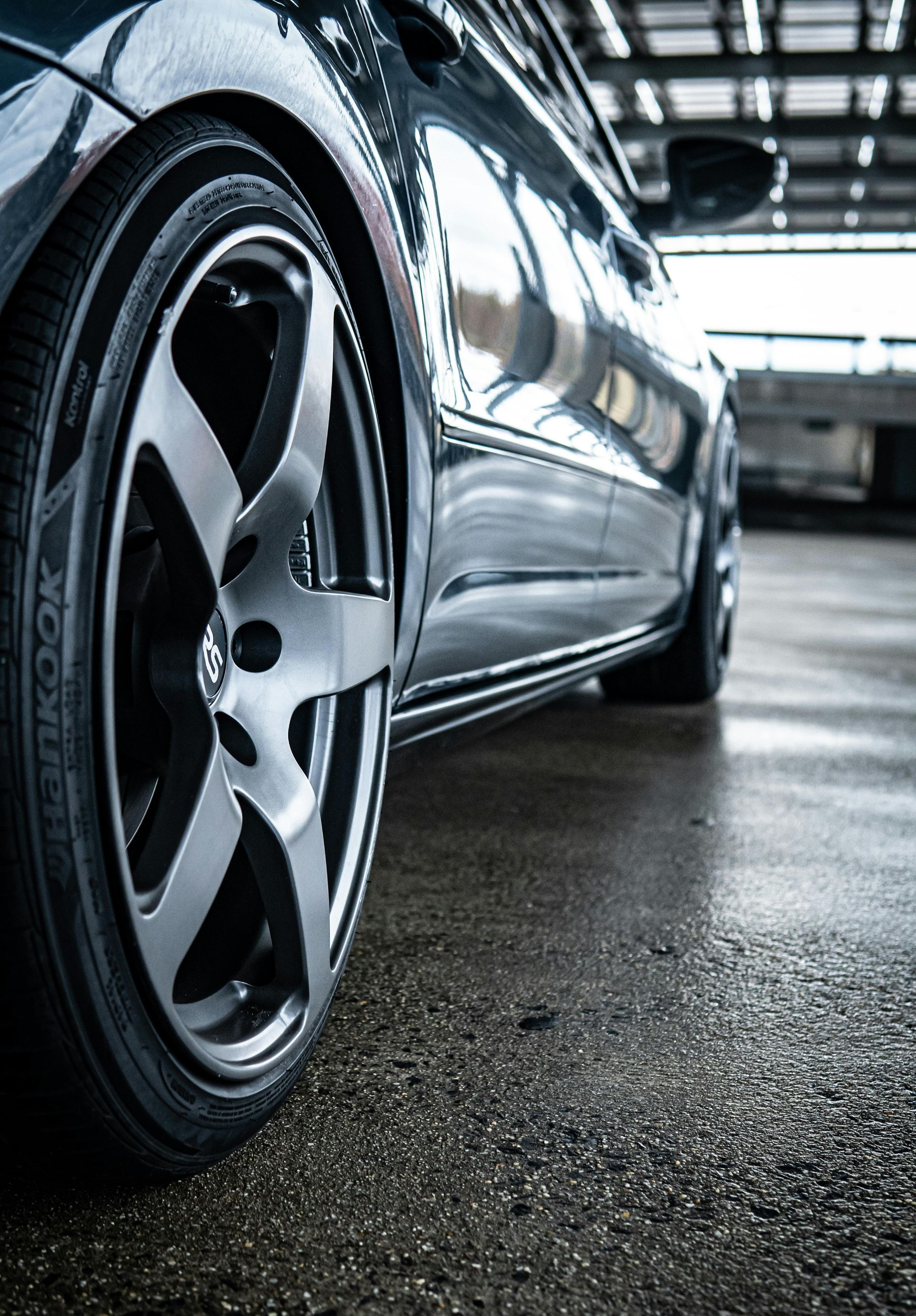 A low-angle, close-up shot of a dark car wheel with a metallic rim parked on a wet, grey concrete surface.