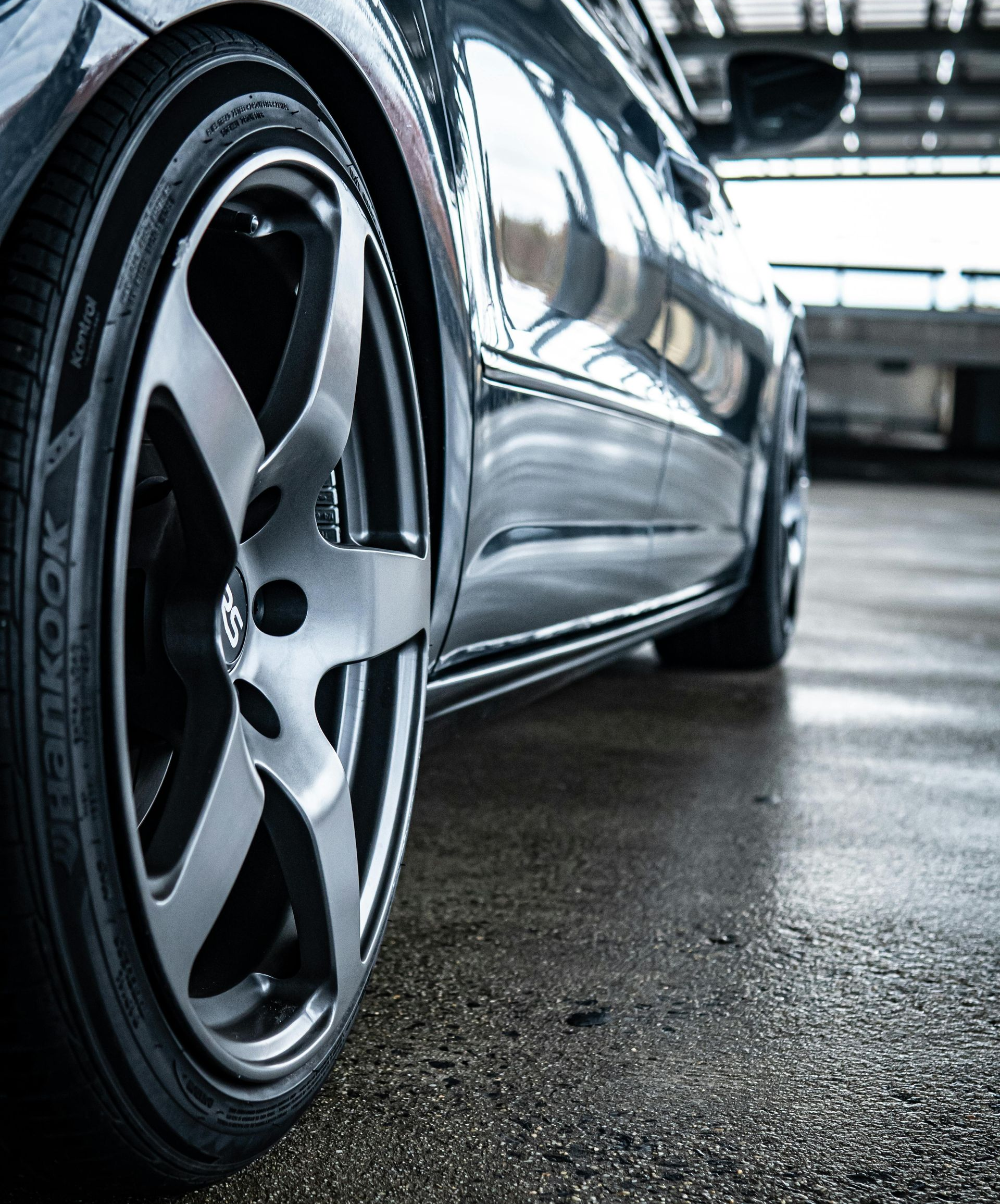 A low-angle, close-up shot of a dark car wheel with a metallic rim parked on a wet, grey concrete surface.
