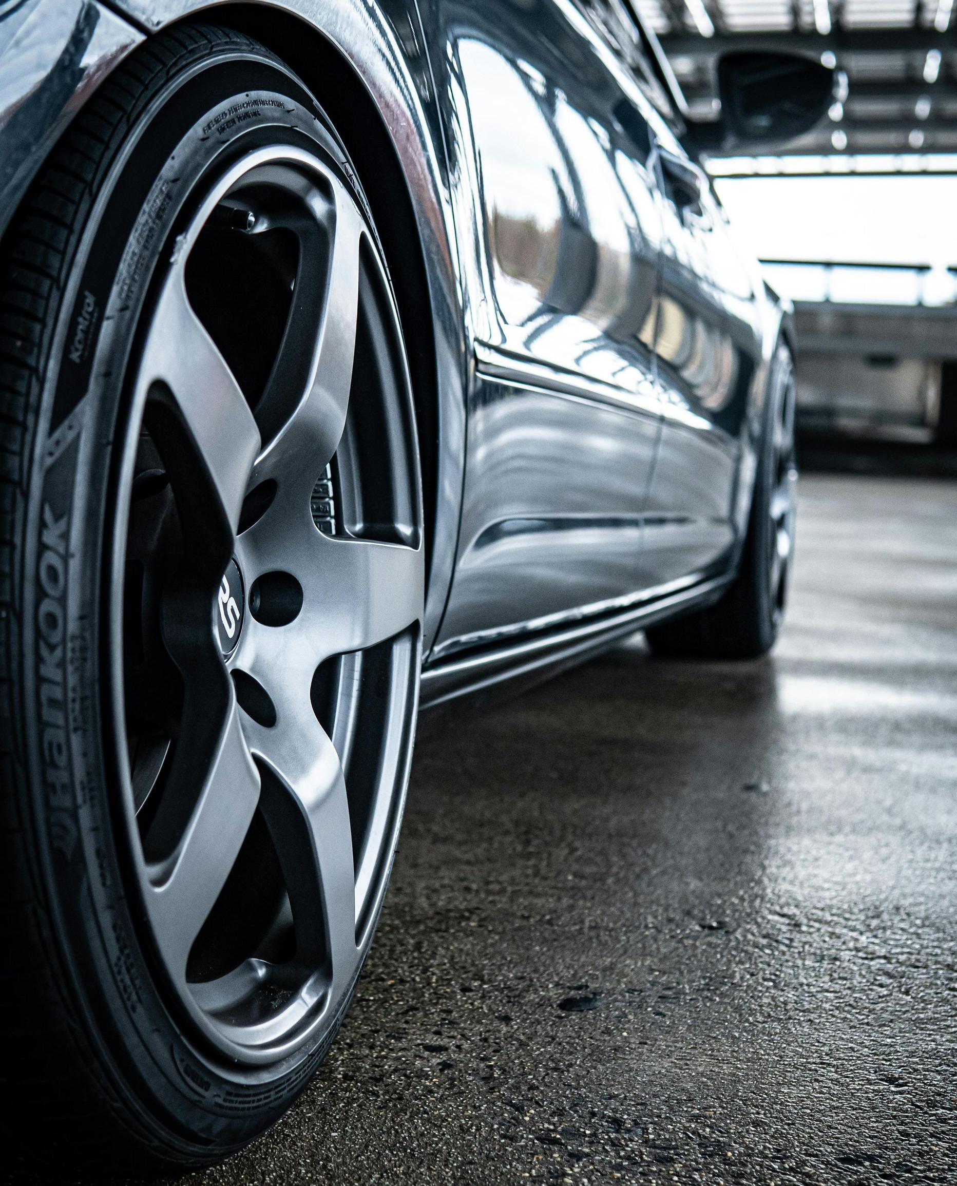 A low-angle, close-up shot of a dark car wheel with a metallic rim parked on a wet, grey concrete surface.