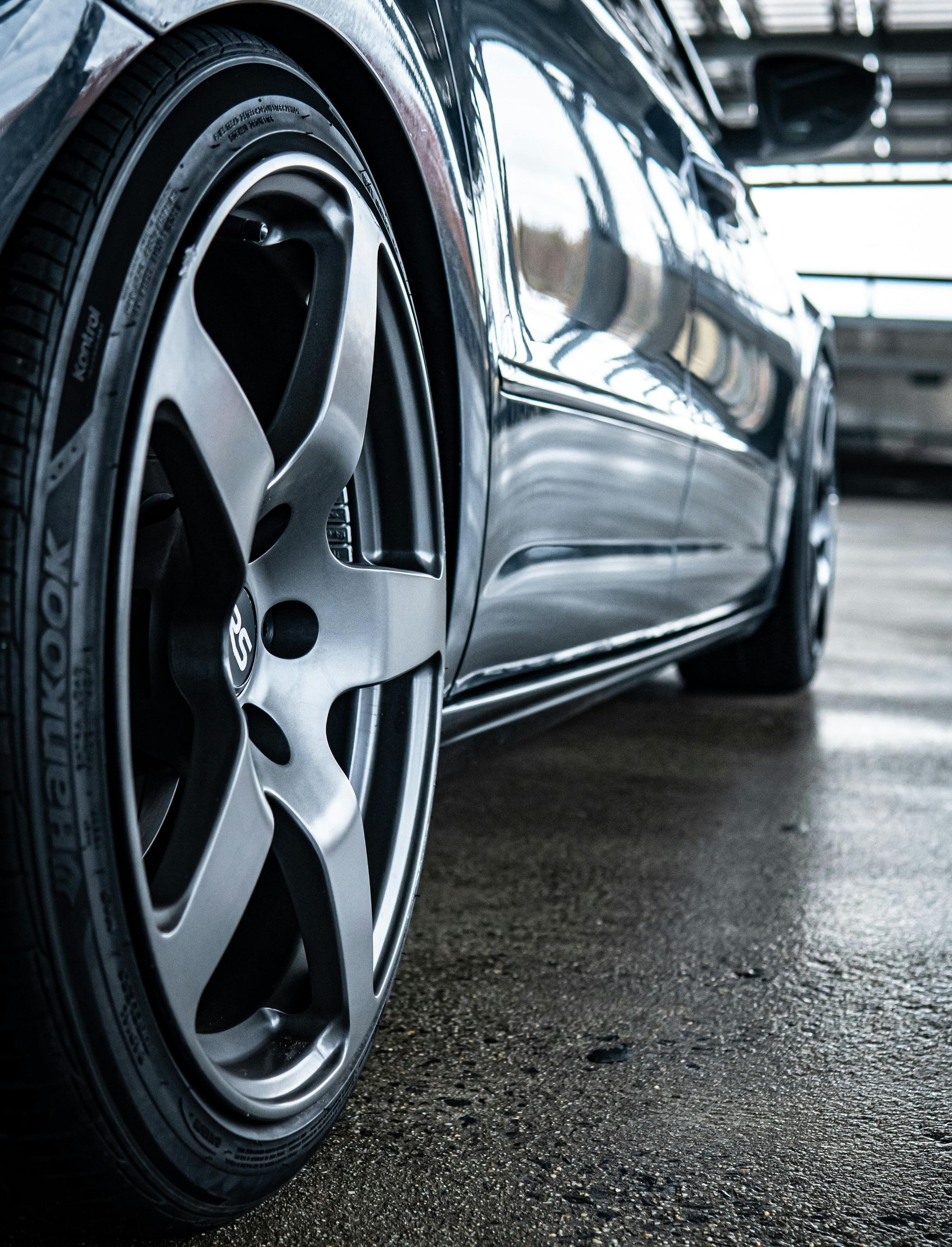 A low-angle, close-up shot of a dark car wheel with a metallic rim parked on a wet, grey concrete surface.