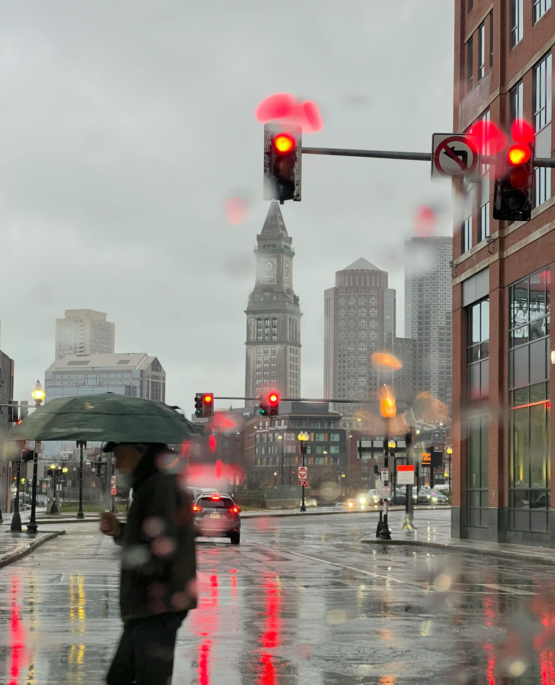 Man with umbrella crossing a rainy street in a city, with red traffic lights and buildings in the background.