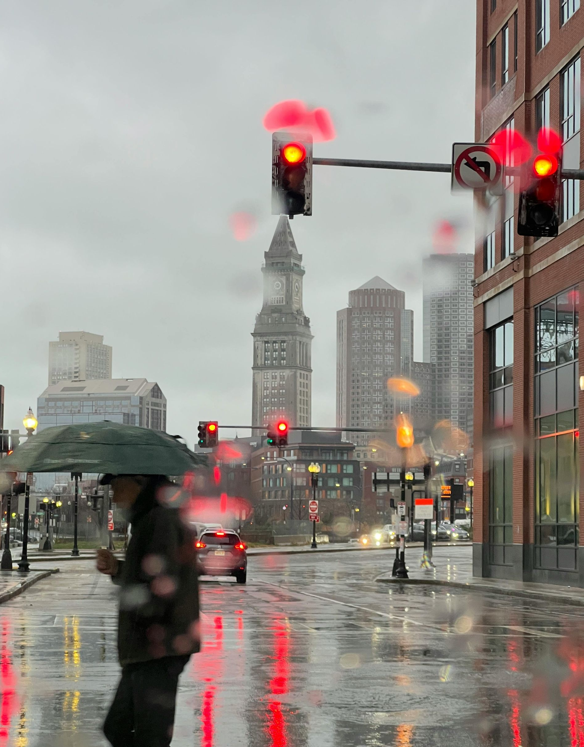 Man with umbrella crossing a rainy street in a city, with red traffic lights and buildings in the background.
