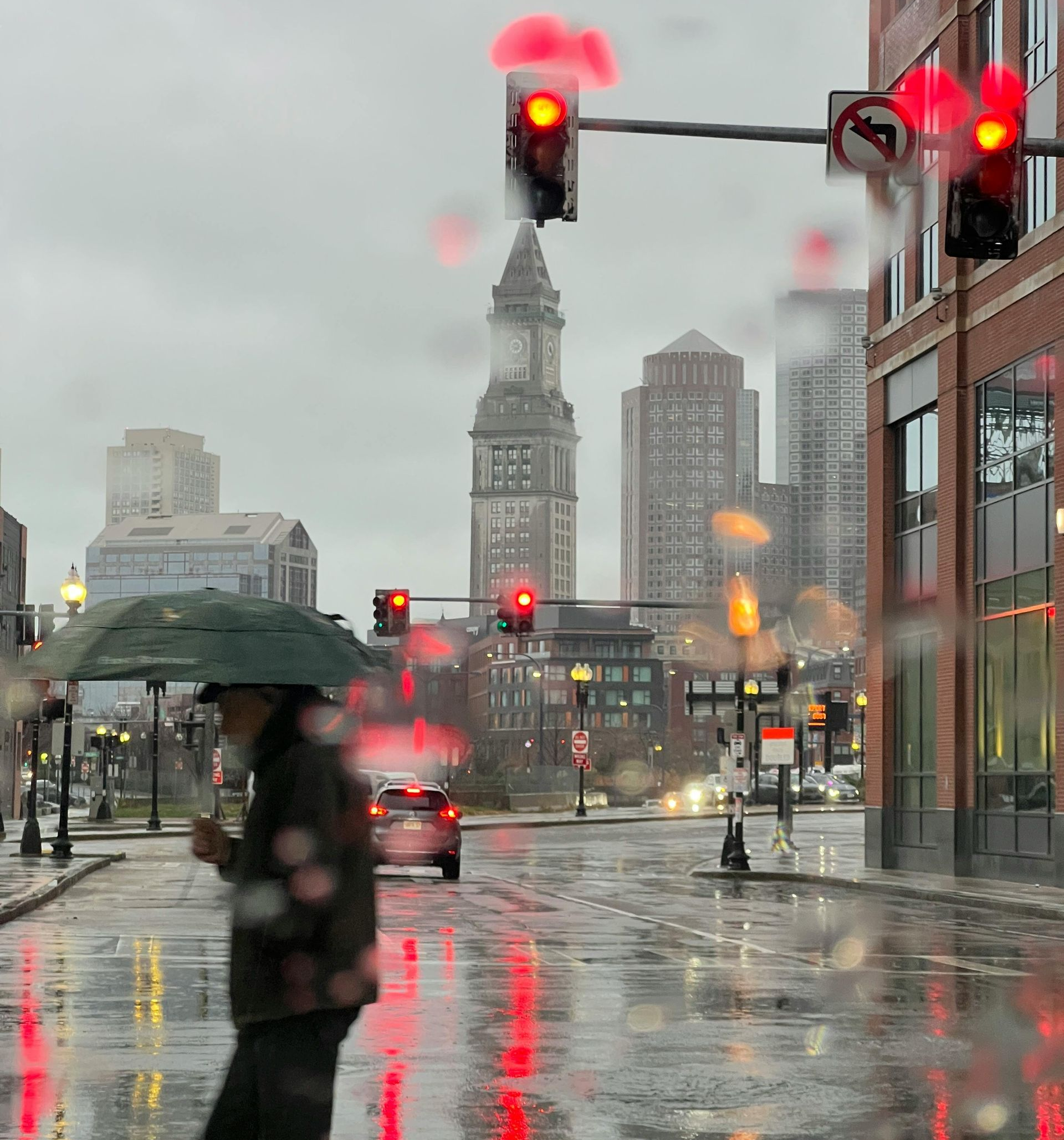 Man with umbrella crossing a rainy street in a city, with red traffic lights and buildings in the background.