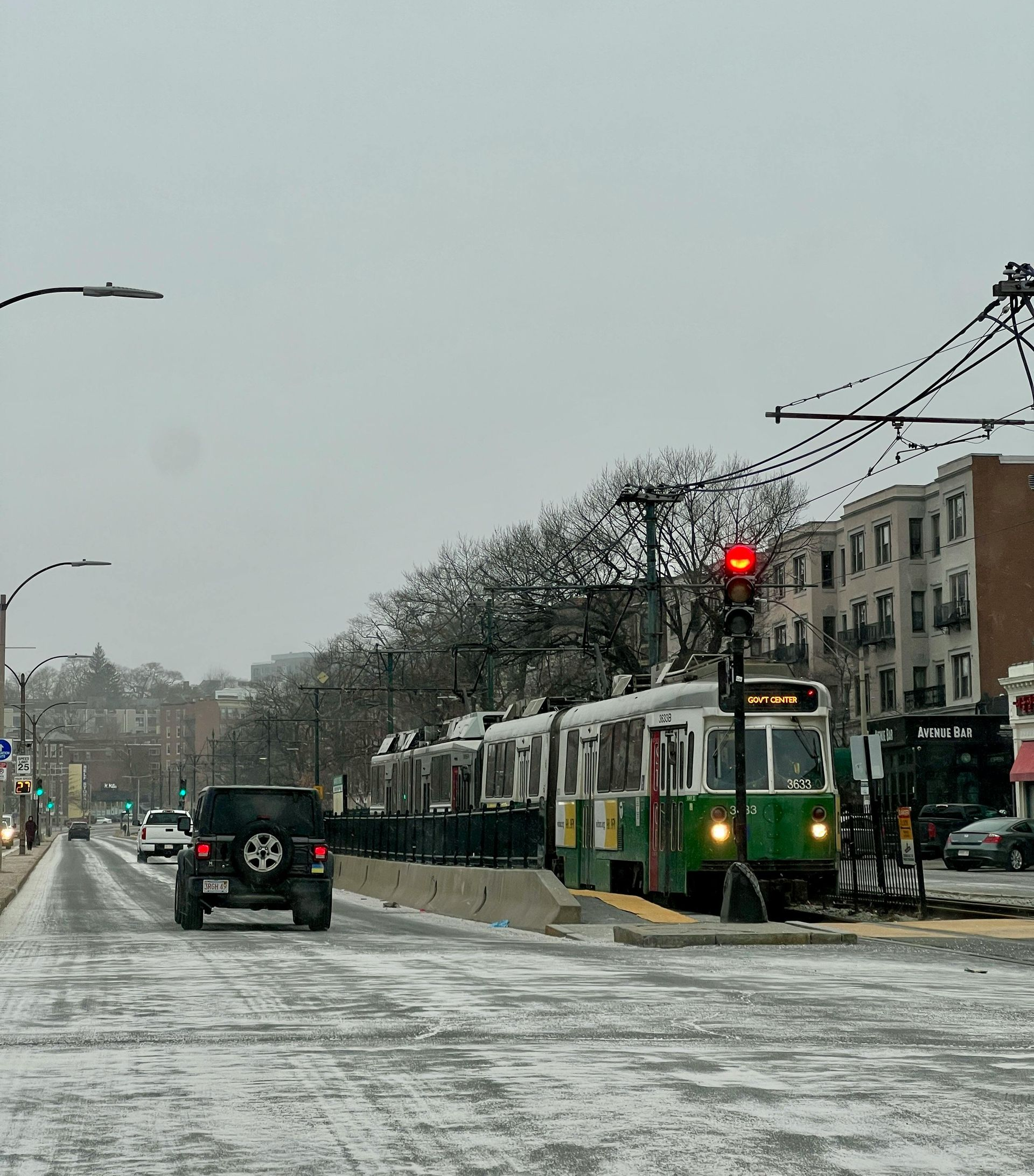 Streetcar stopped at a station on a snow-covered street; cars and buildings in the background.