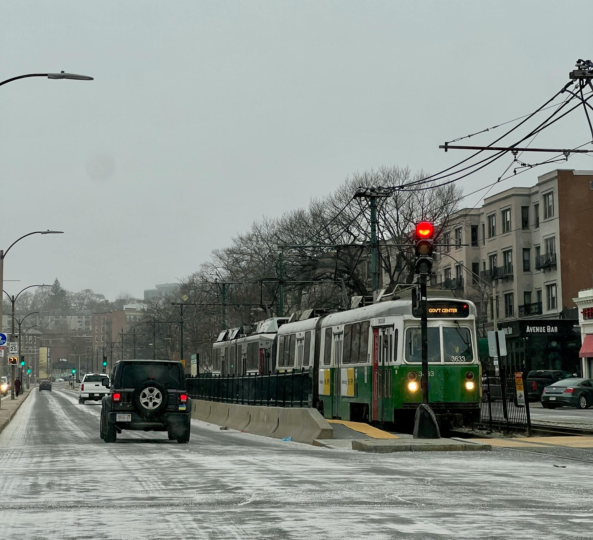 Streetcar stopped at a station on a snow-covered street; cars and buildings in the background.