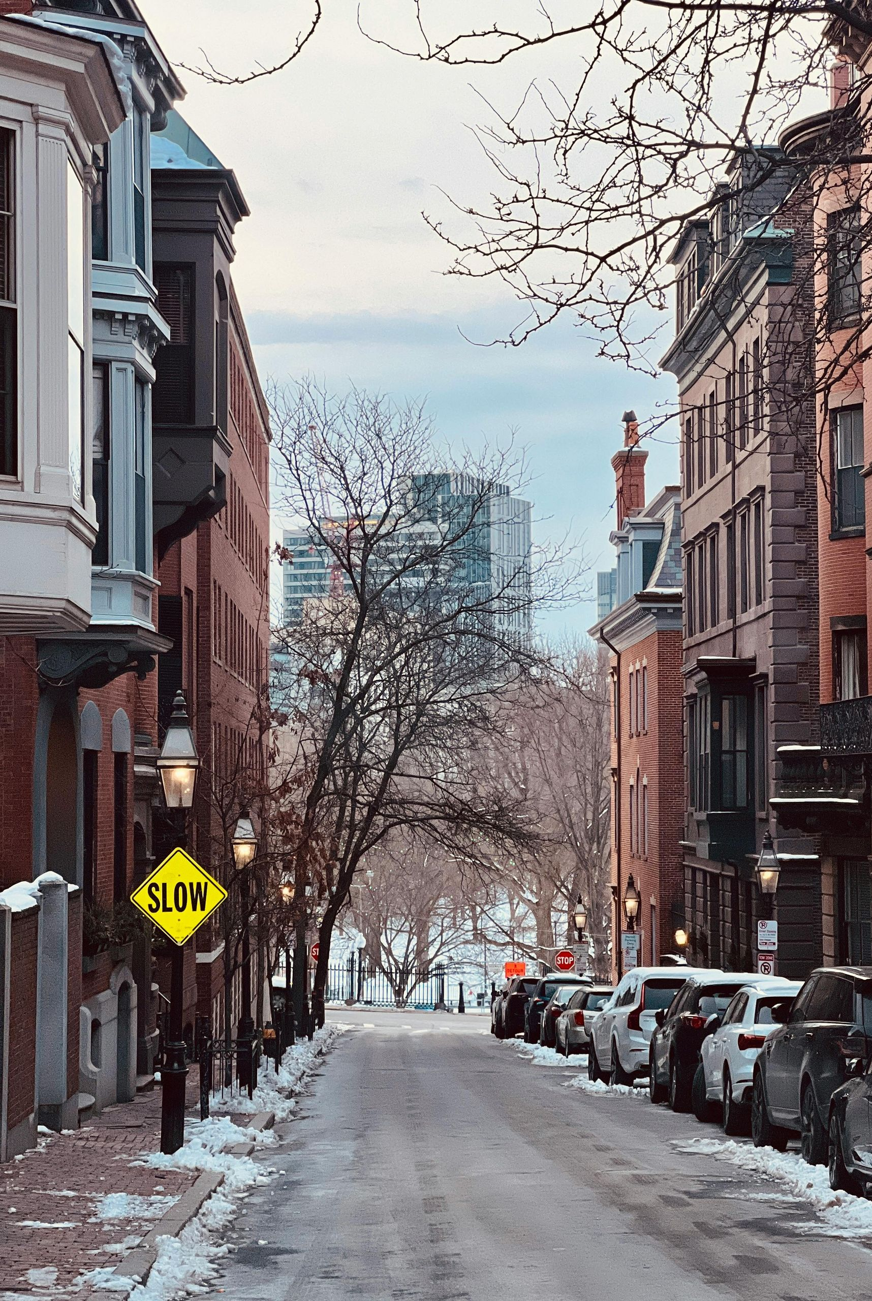 Snowy street lined with brownstone buildings, cars parked on the side. A