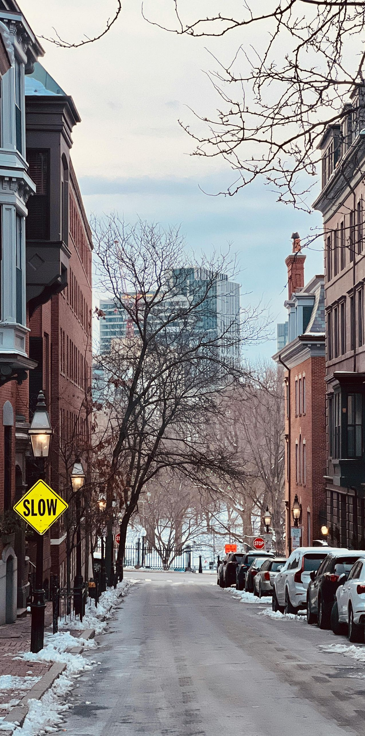 Snowy street lined with brownstone buildings, cars parked on the side.  A 