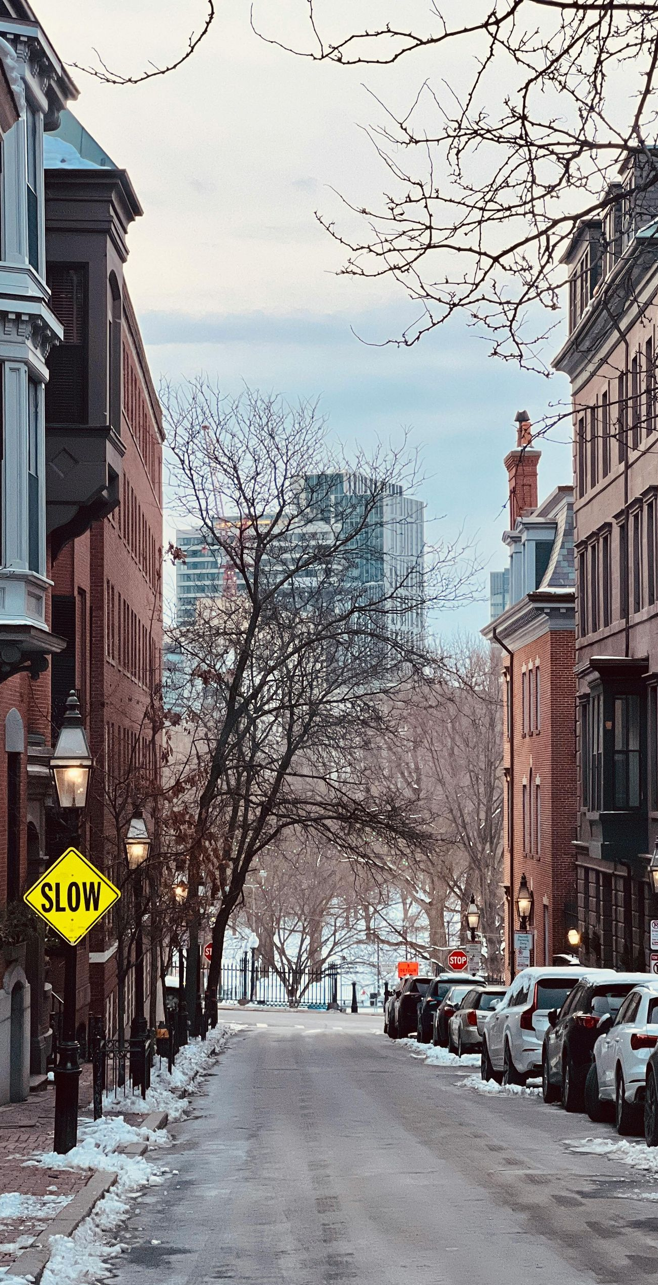 Snowy street lined with brownstone buildings, cars parked on the side. A