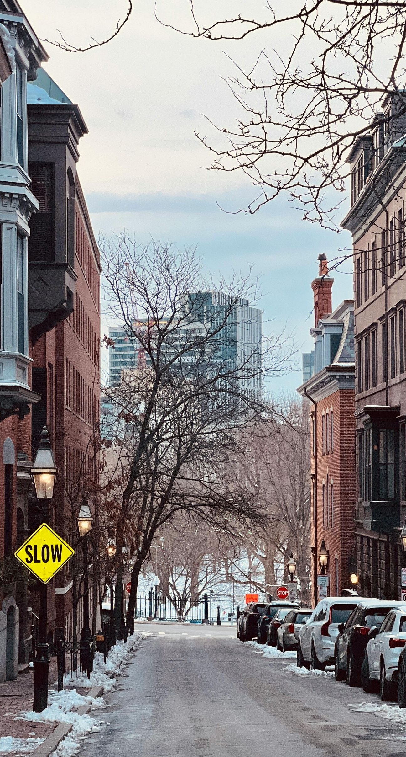 Snowy street lined with brownstone buildings, cars parked on the side. A