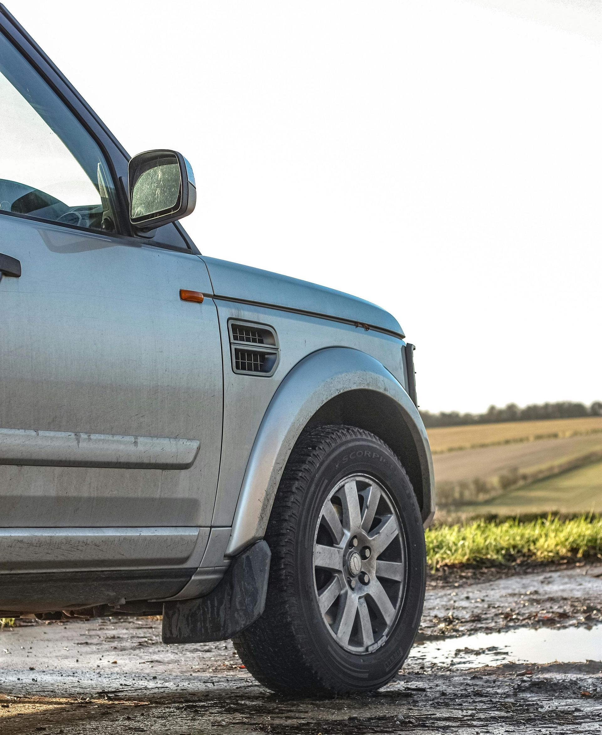 Silver Land Rover SUV on a muddy track, near a field. Partially visible, sunlit.