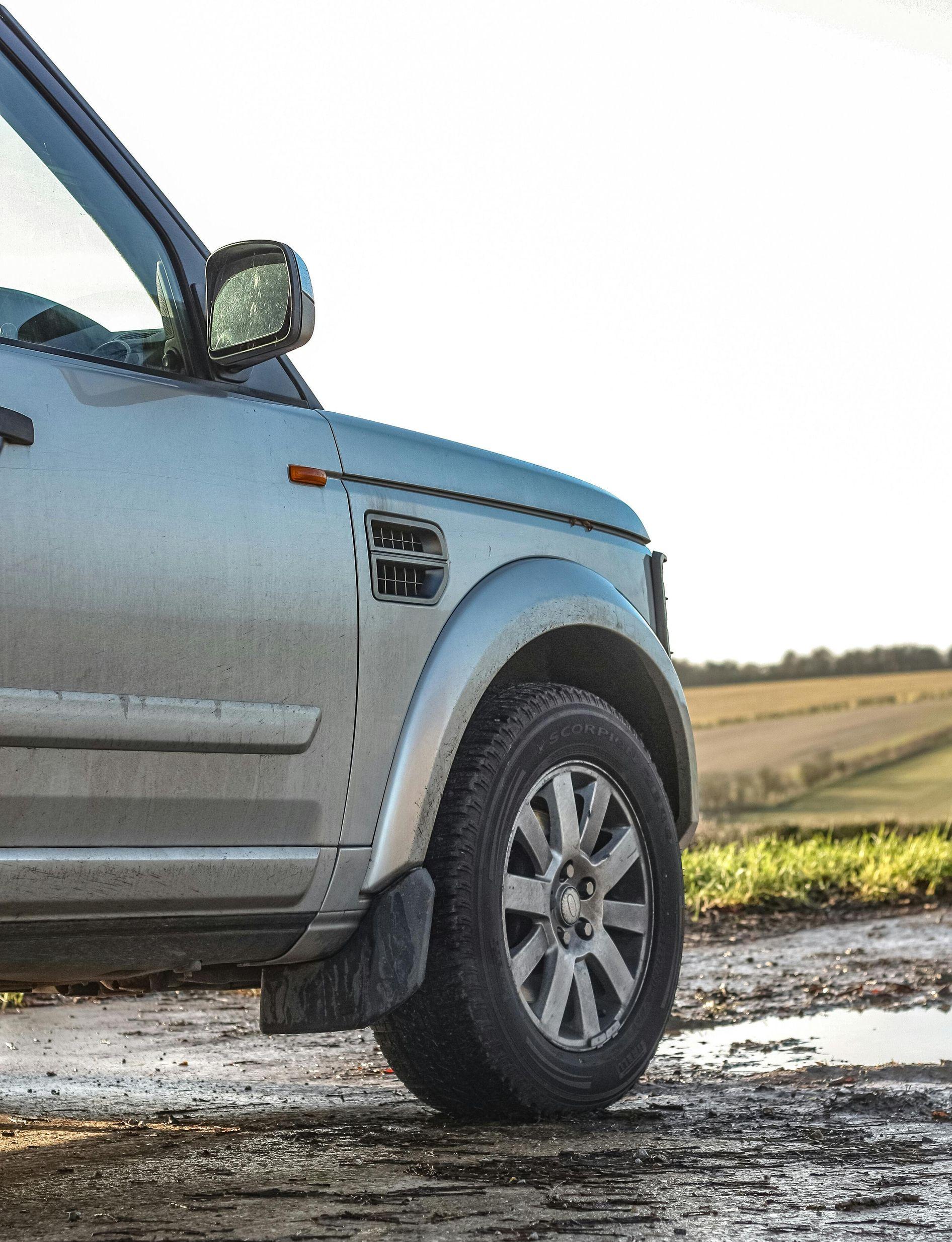 Silver Land Rover SUV on a muddy track, near a field. Partially visible, sunlit.