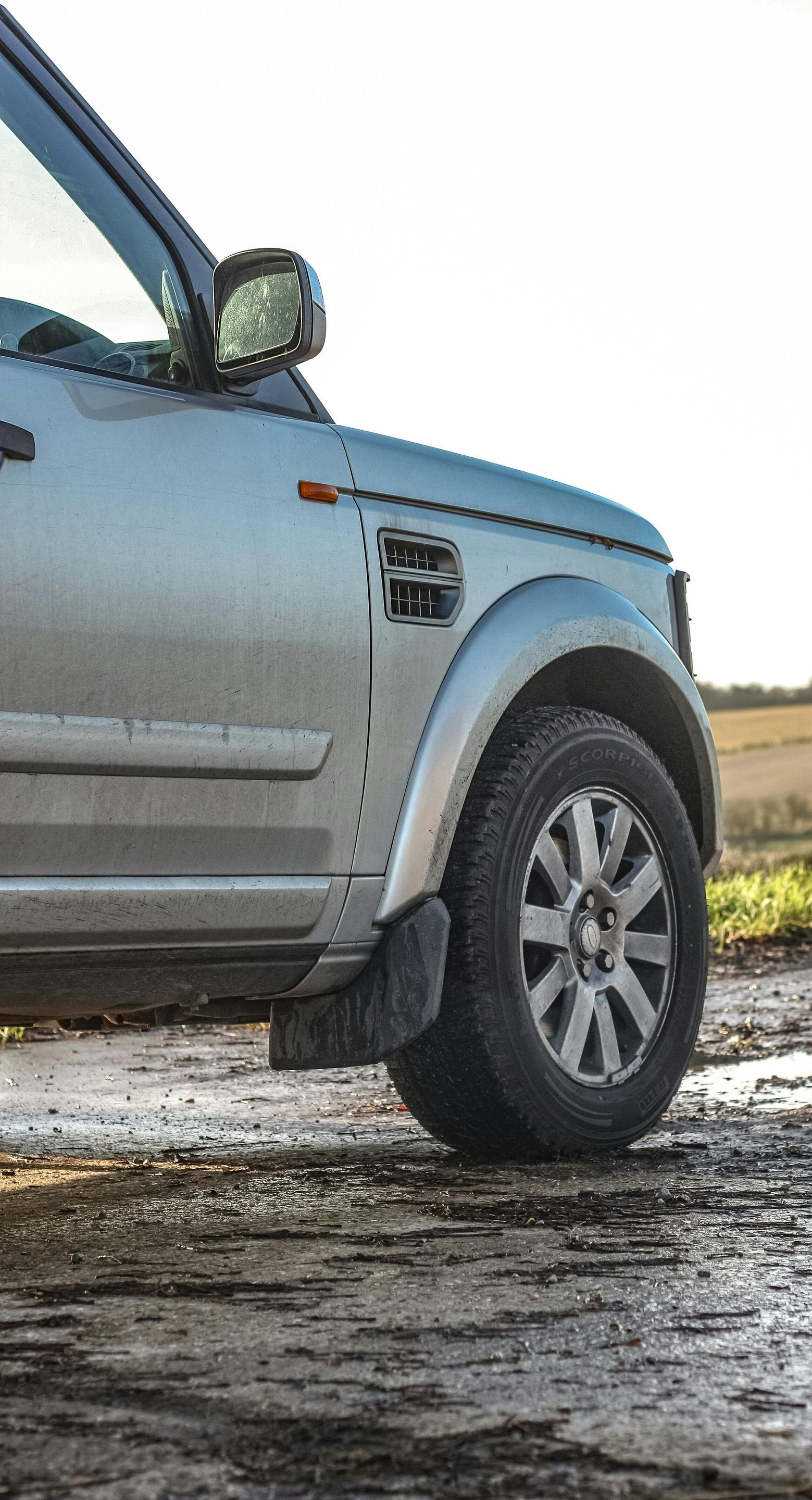 Silver Land Rover SUV on a muddy track, near a field. Partially visible, sunlit.