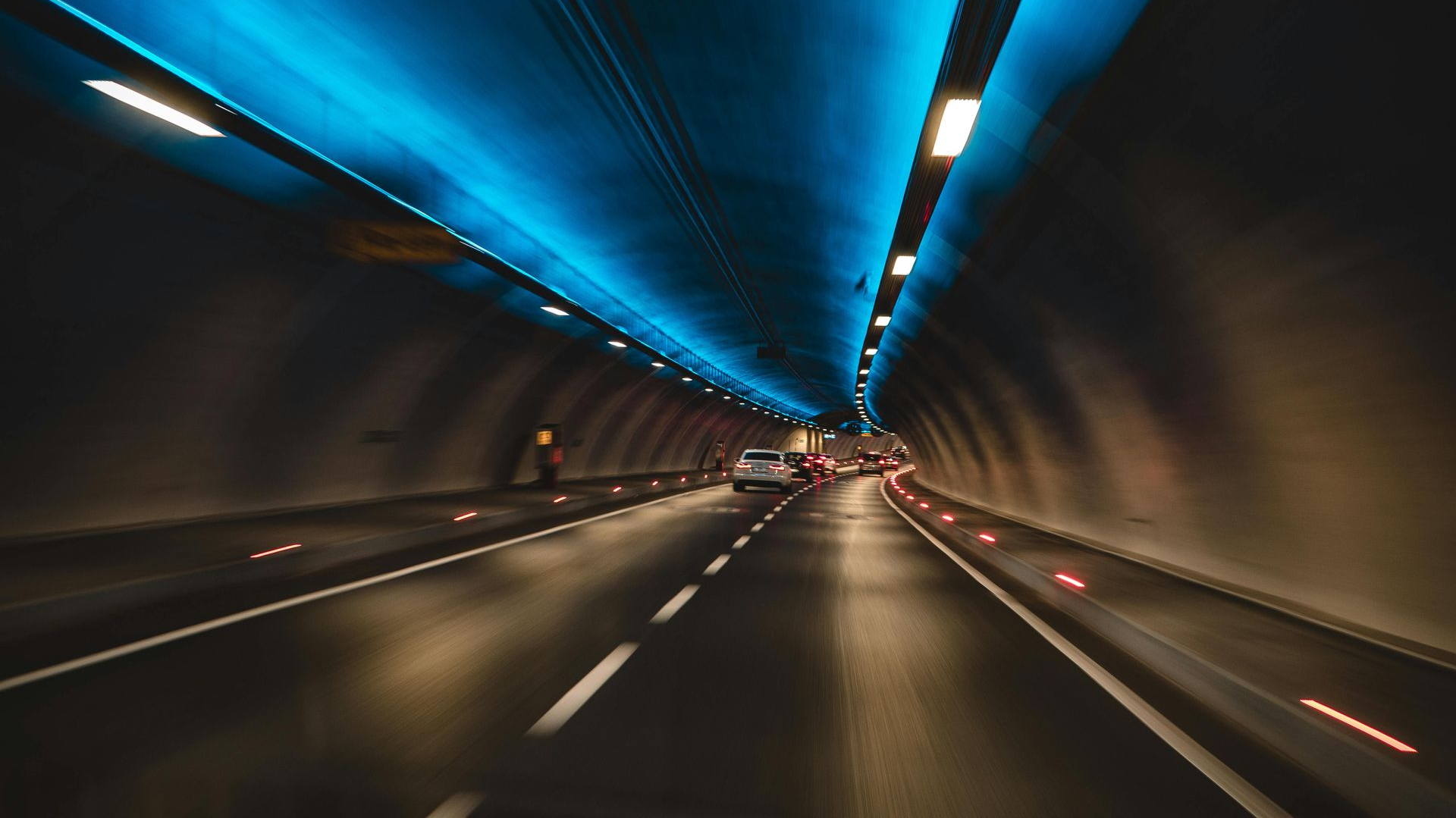 Inside a tunnel, cars driving toward the light. Blue ceiling lights and road markers glow.