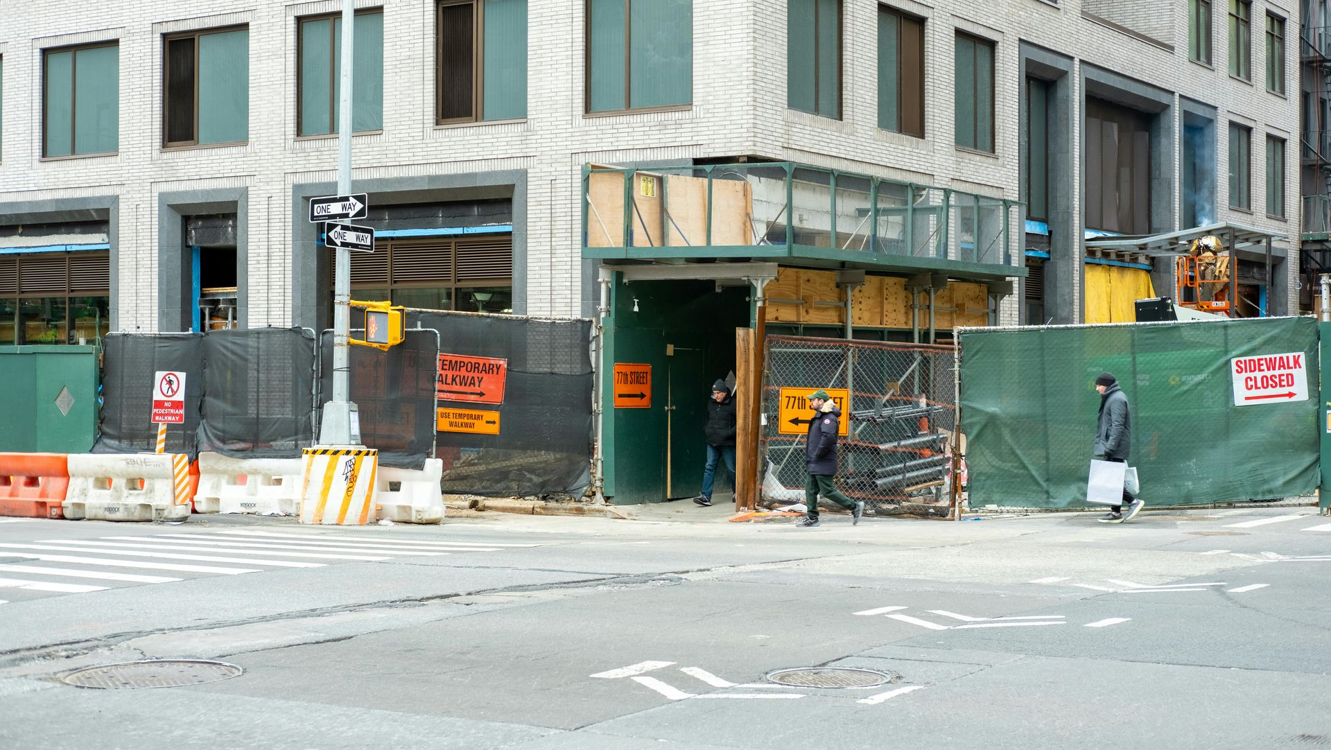 Street-level view of a construction site beside a stone building, with green barriers and traffic cones at the corner.