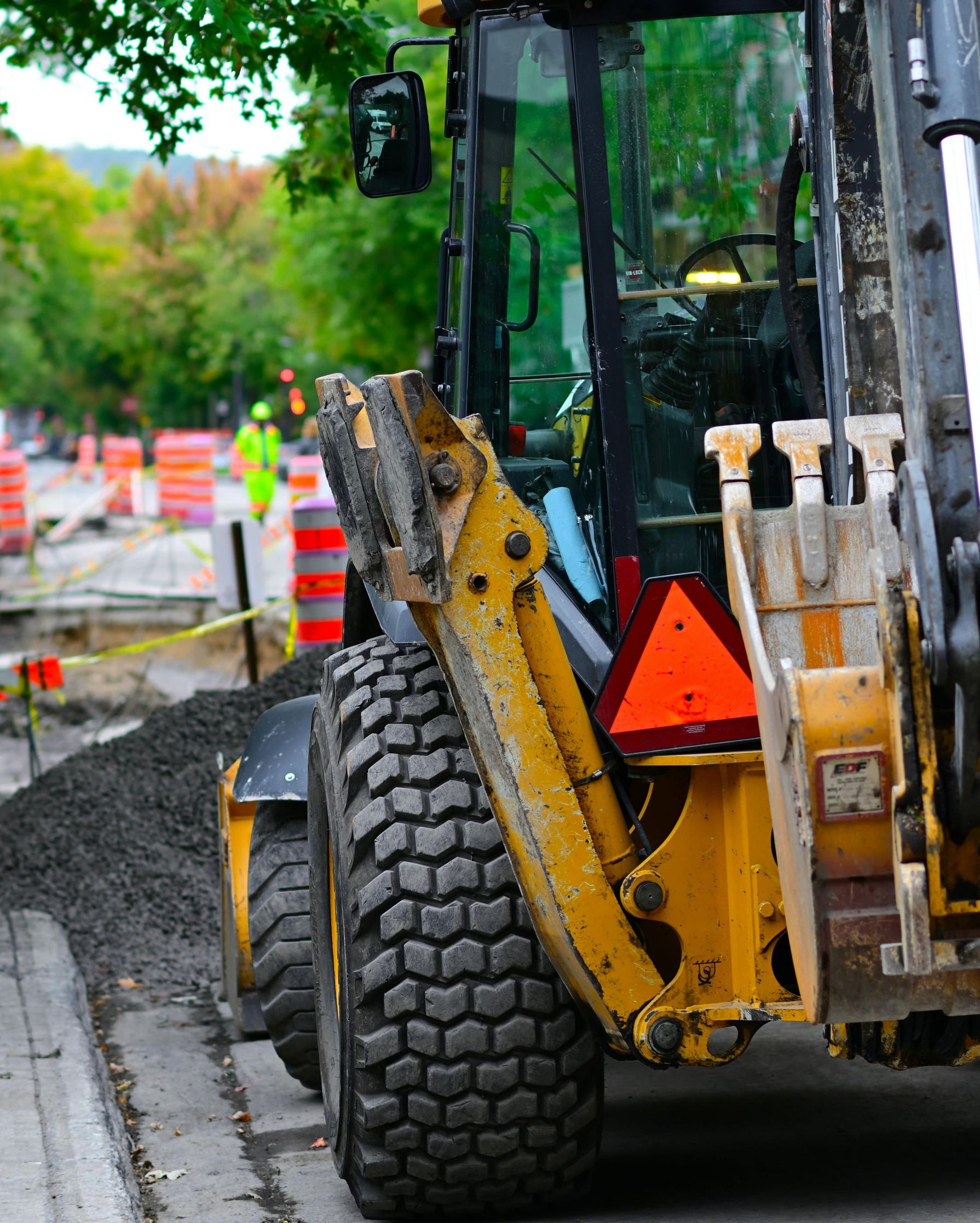 Yellow backhoe on a wet roadside beside a tree and traffic cones, with a large tire in the foreground