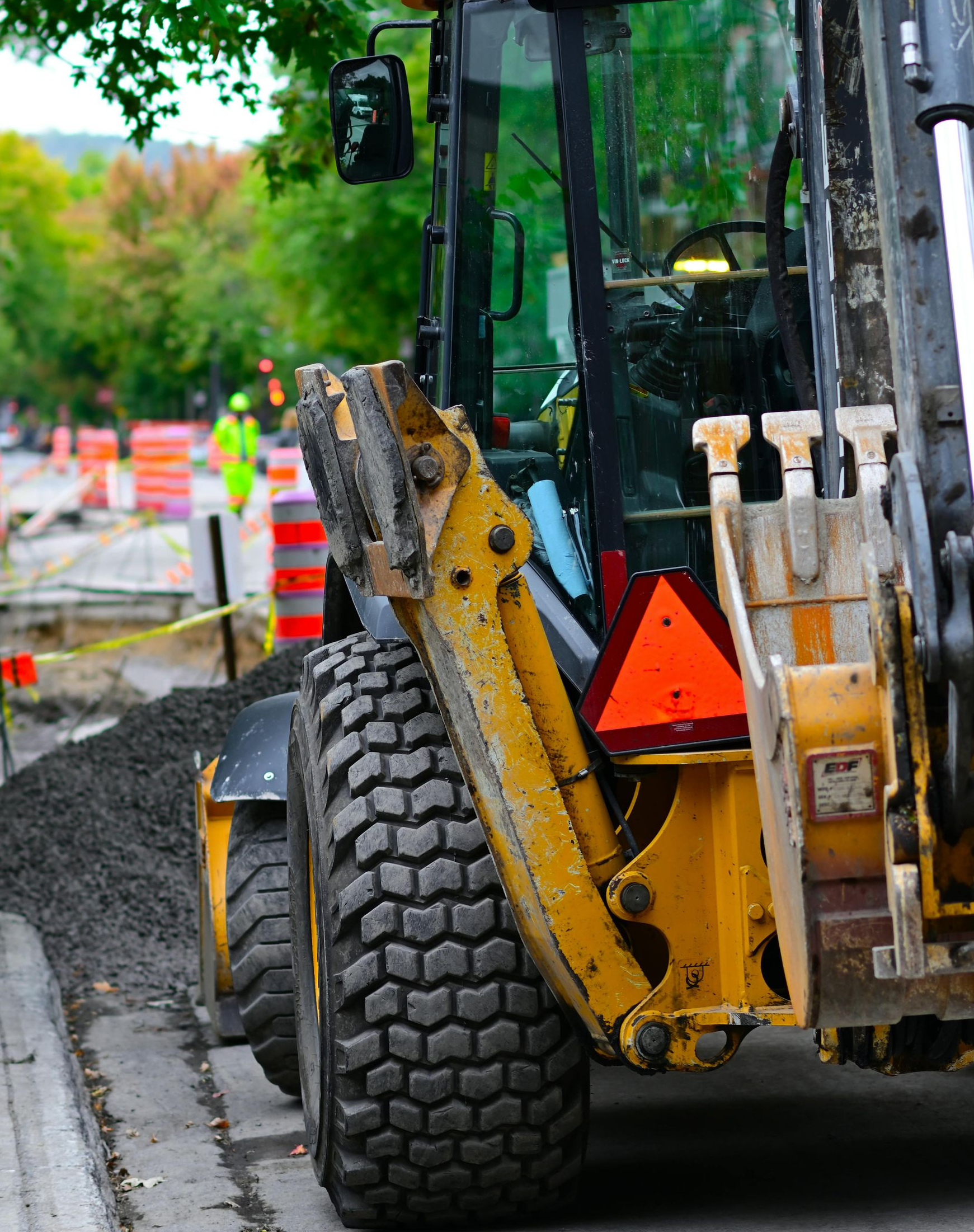 Yellow backhoe on a wet roadside beside a tree and traffic cones, with a large tire in the foreground