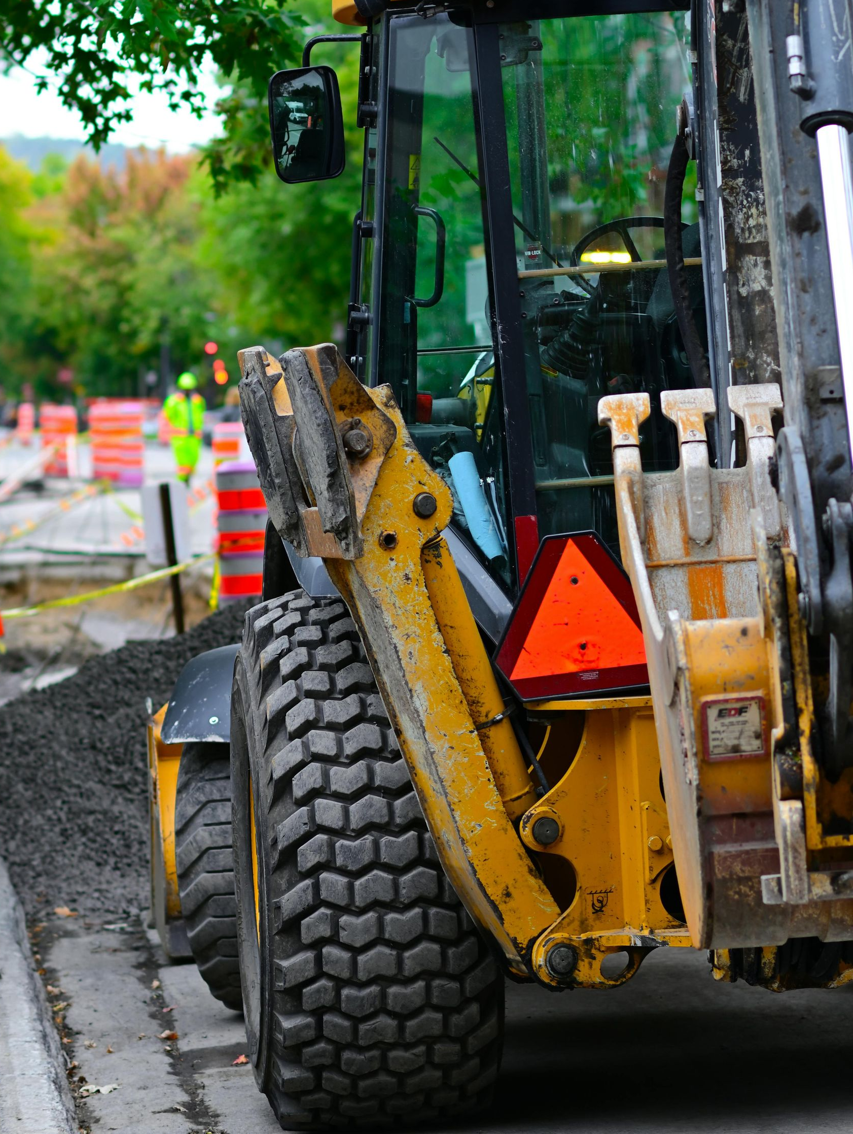 Yellow backhoe on a wet roadside beside a tree and traffic cones, with a large tire in the foreground