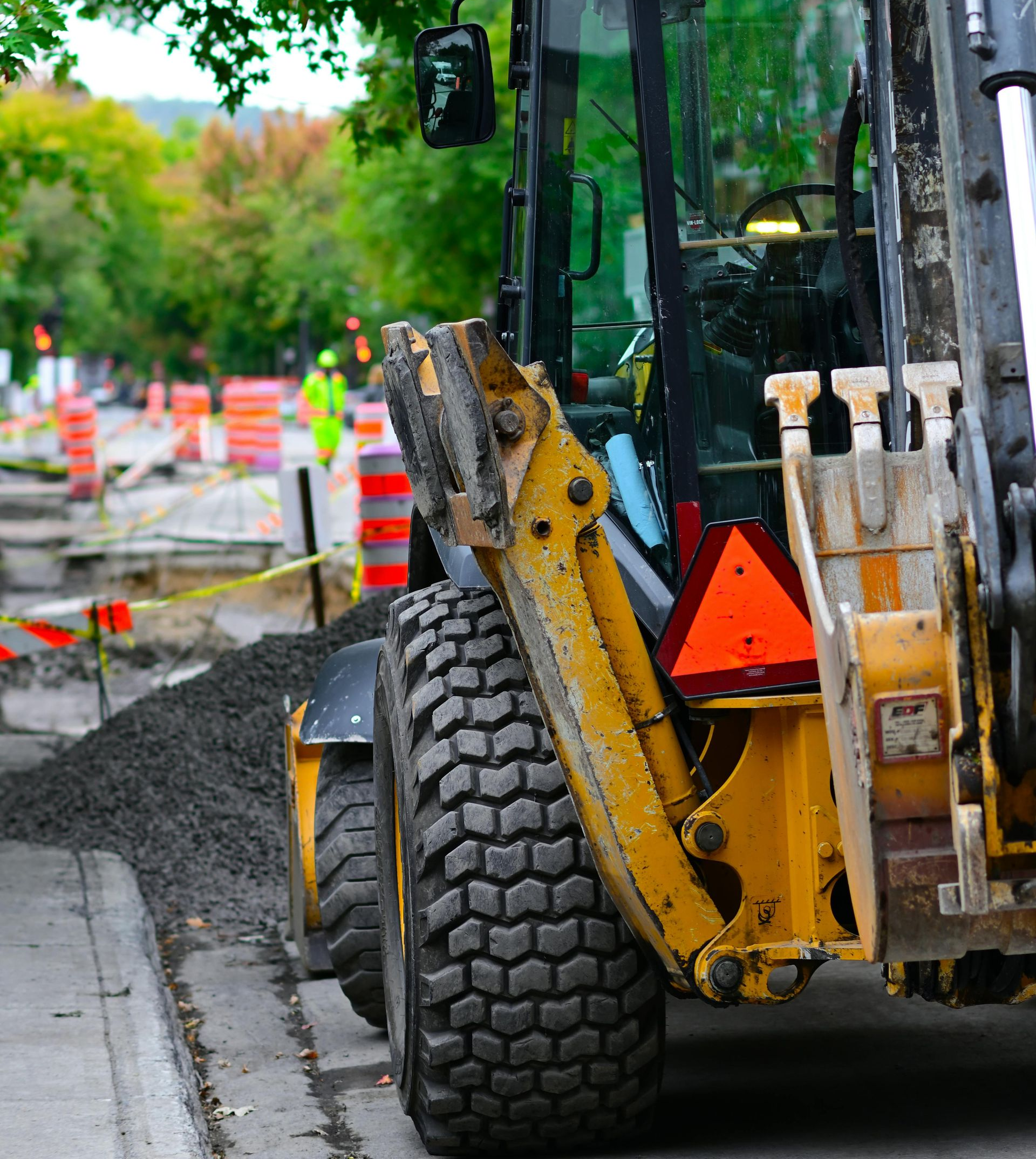 Yellow backhoe on a wet roadside beside a tree and traffic cones, with a large tire in the foreground