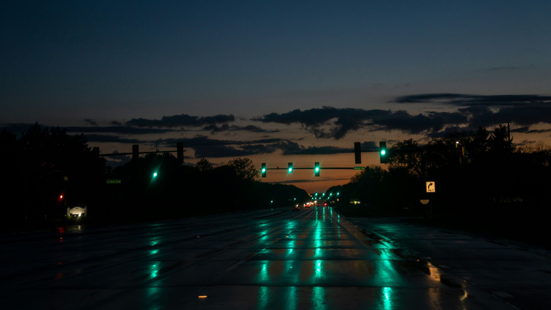 Wet road at dusk with green traffic lights reflecting on the pavement under a cloudy sky