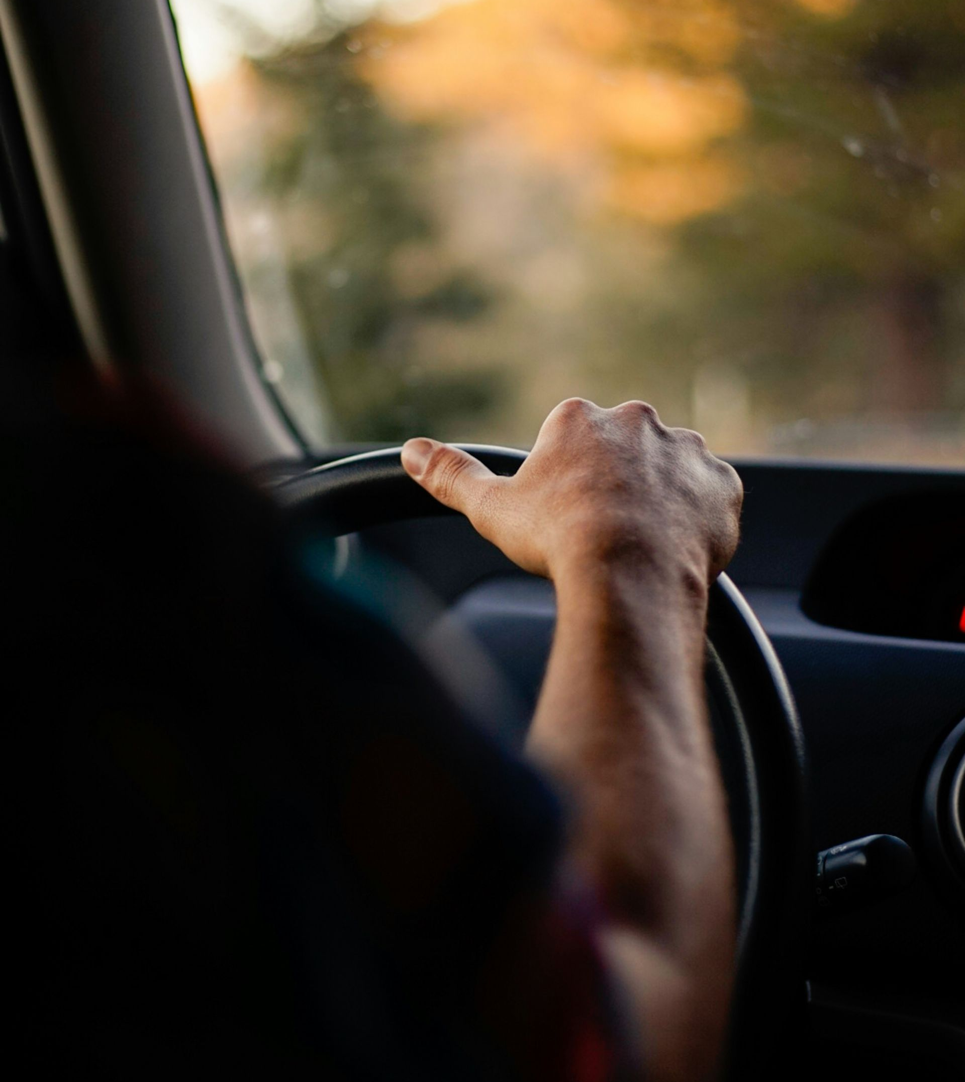 Driver’s hand on steering wheel in a car, with a blurred sunset view ahead.